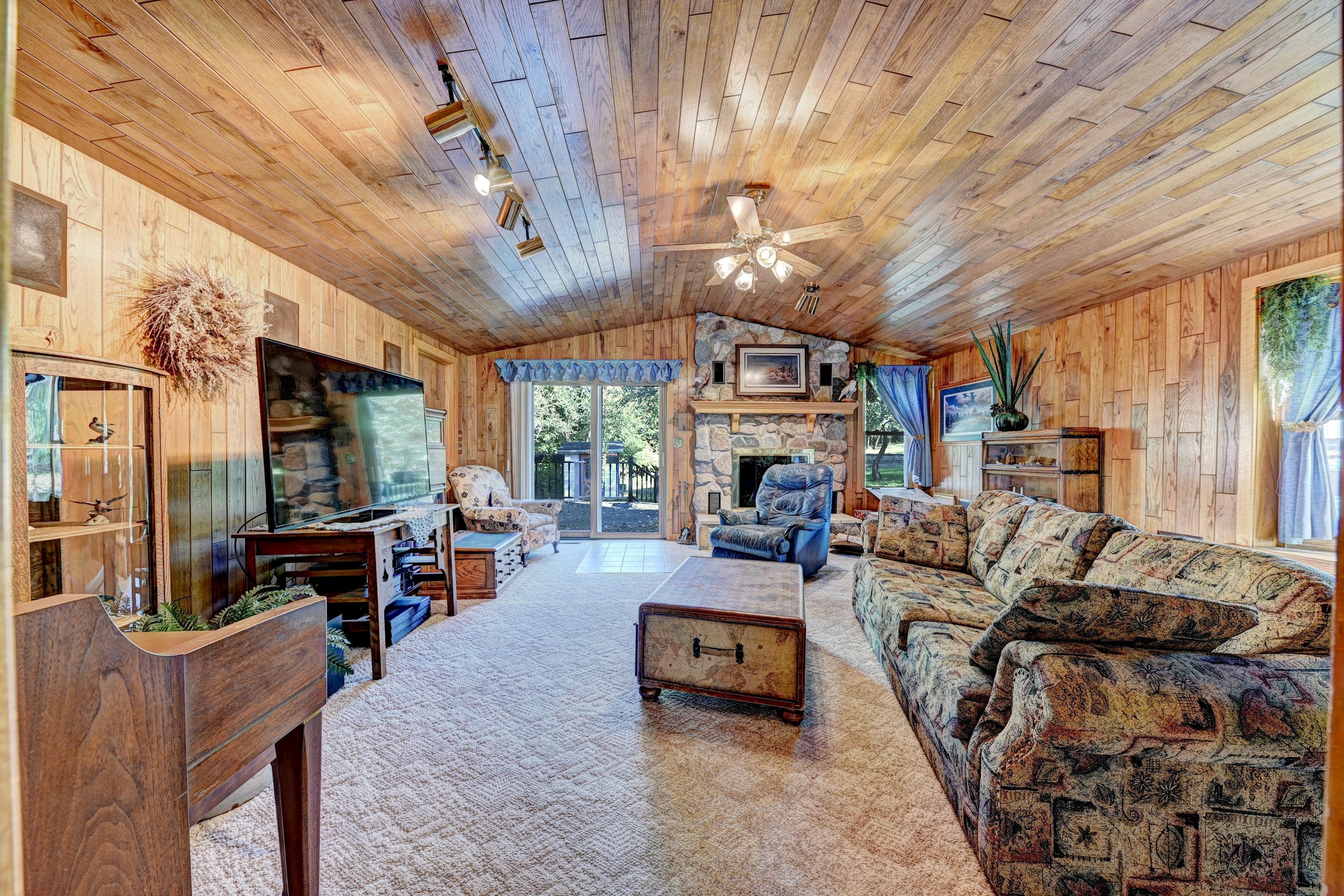 Living room with wood-paneled ceiling and walls, a stone fireplace, large sliding glass doors, and various sofas and chairs.