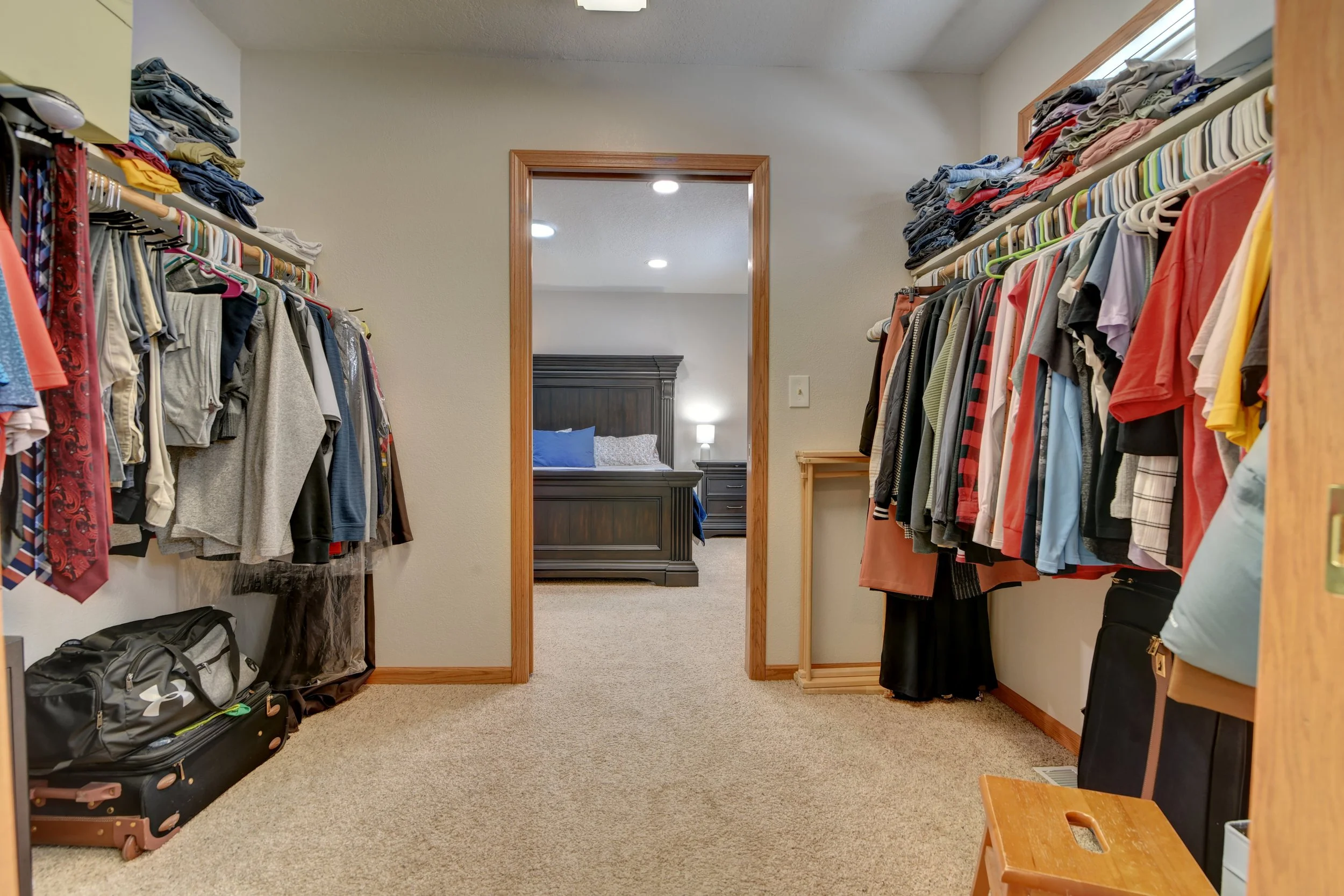 A walk-in closet with organized clothing on racks and shelves, leading to a bedroom with a dark wood bed and nightstand with a lamp.