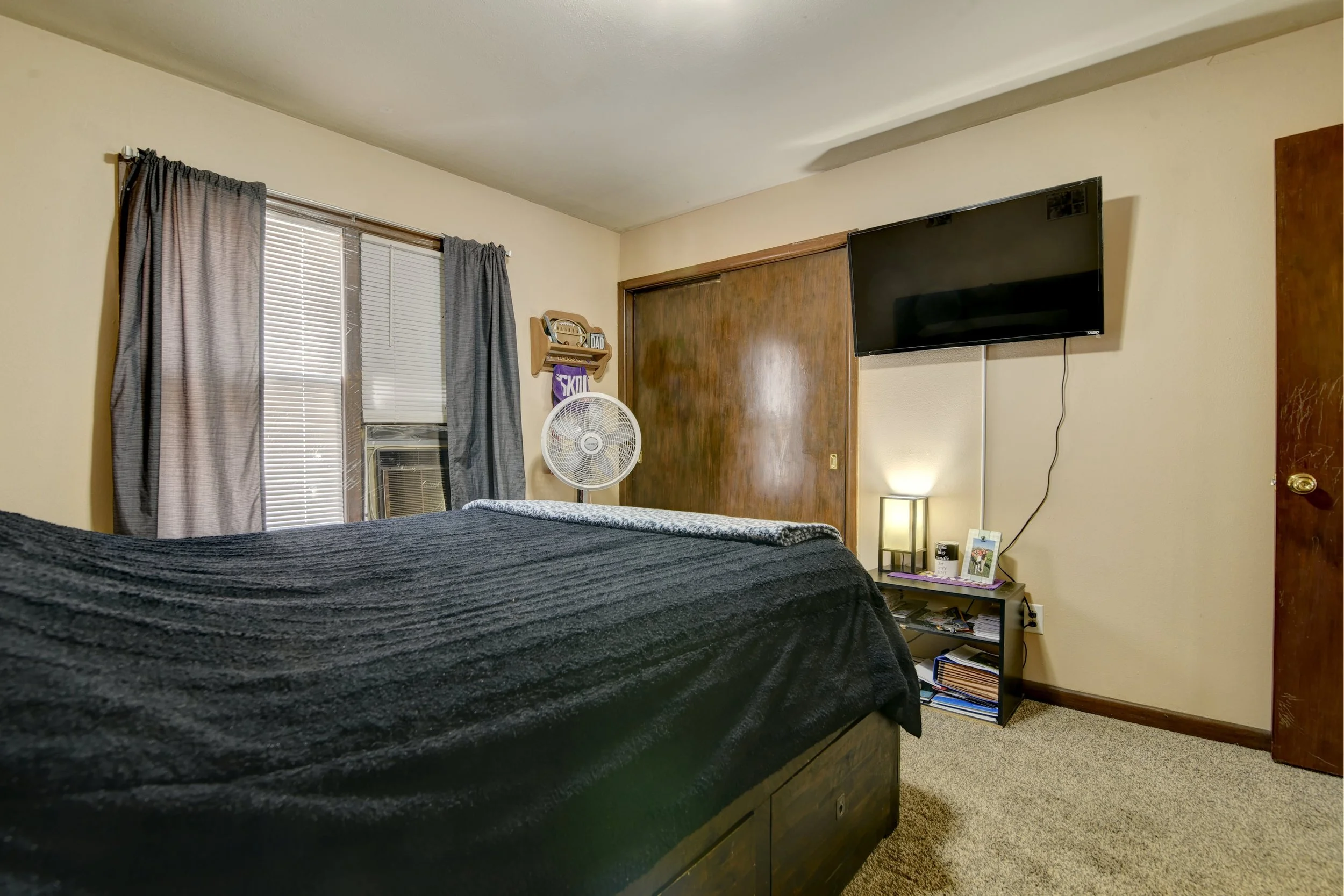 A bedroom with a bed covered in a dark blanket, a TV mounted on the wall, a small nightstand with books, a fan, and framed photos, a window with blinds and curtains, a wooden door, and an air conditioning unit.
