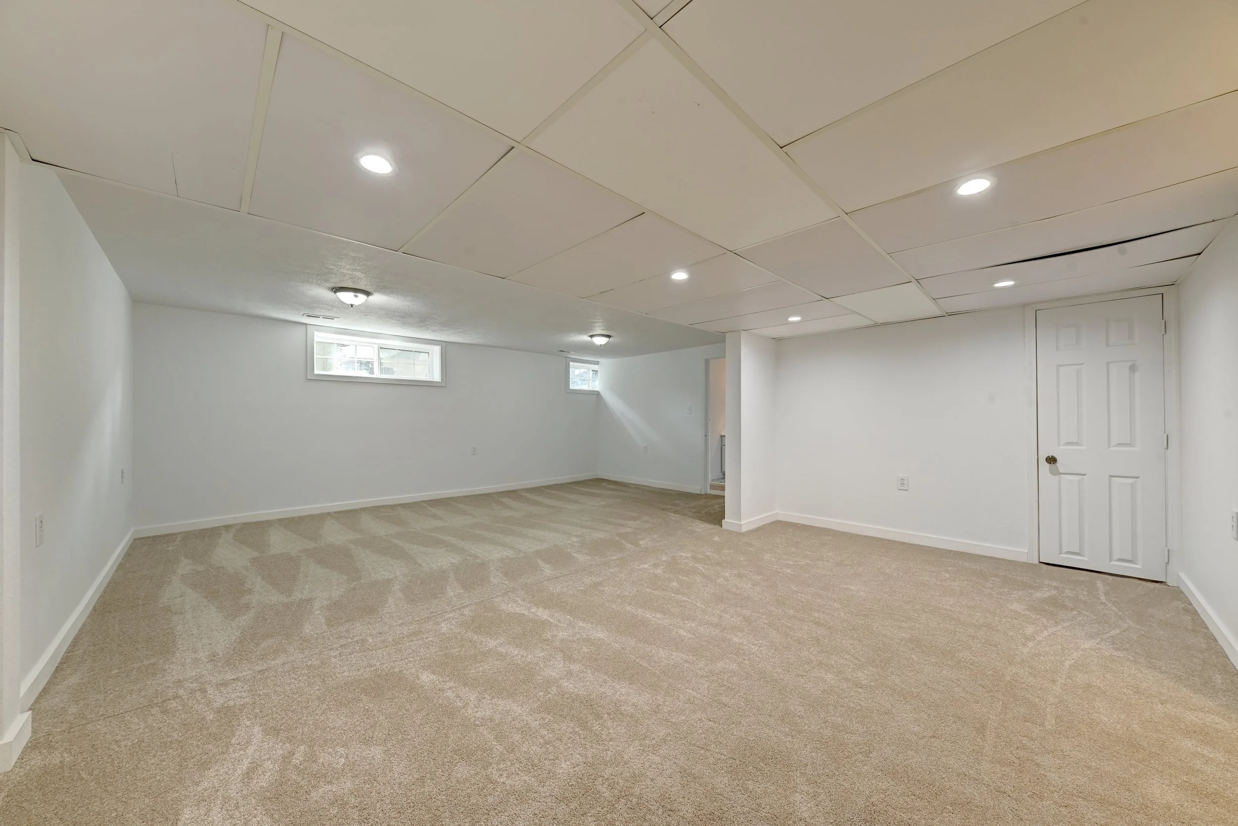 Empty finished basement with beige carpet, white walls, a white ceiling with recessed lighting, small high windows, and a white door.