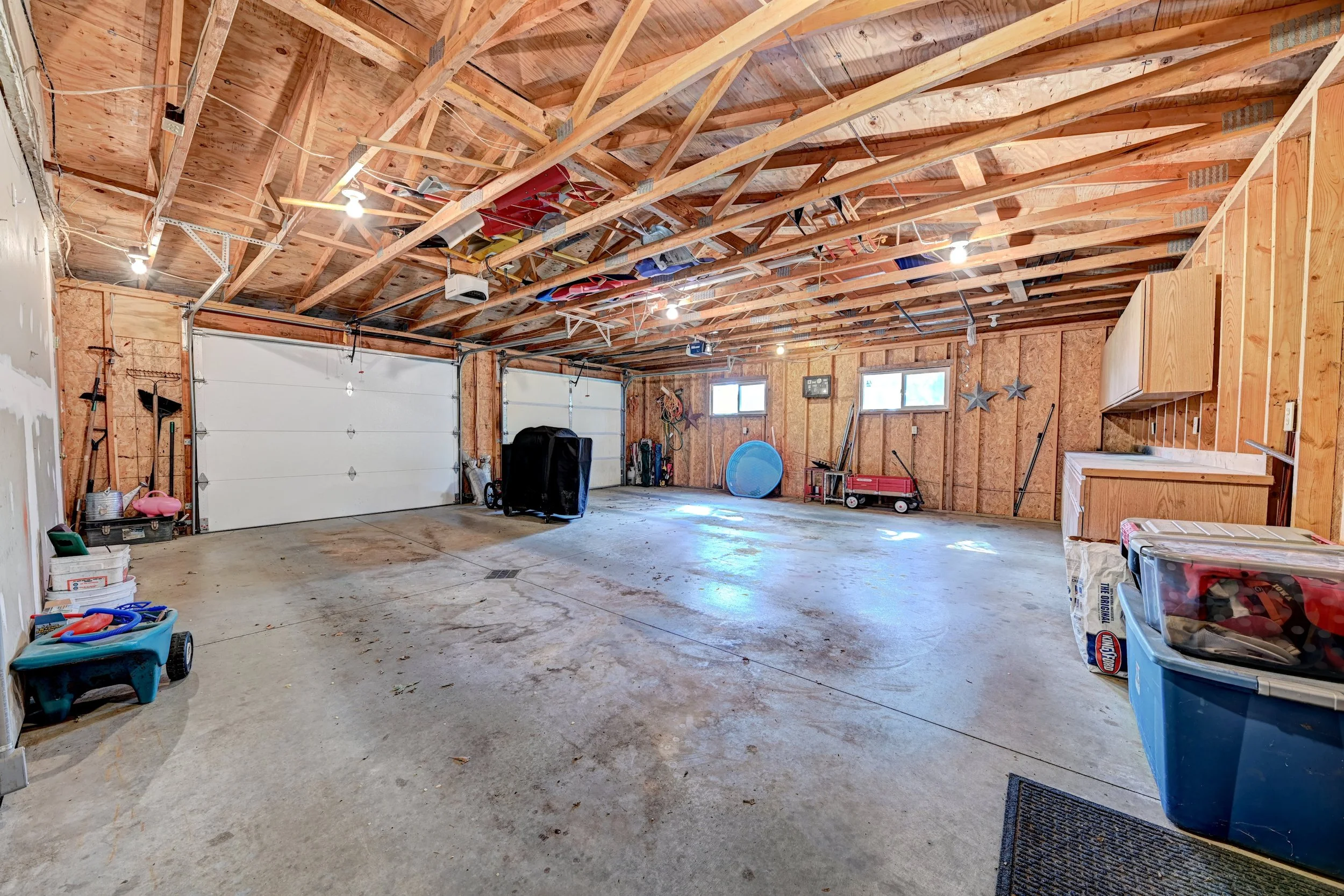 Empty garage with unfinished wood ceiling, two storage garage doors, and various tools and items stored along the walls and floor.