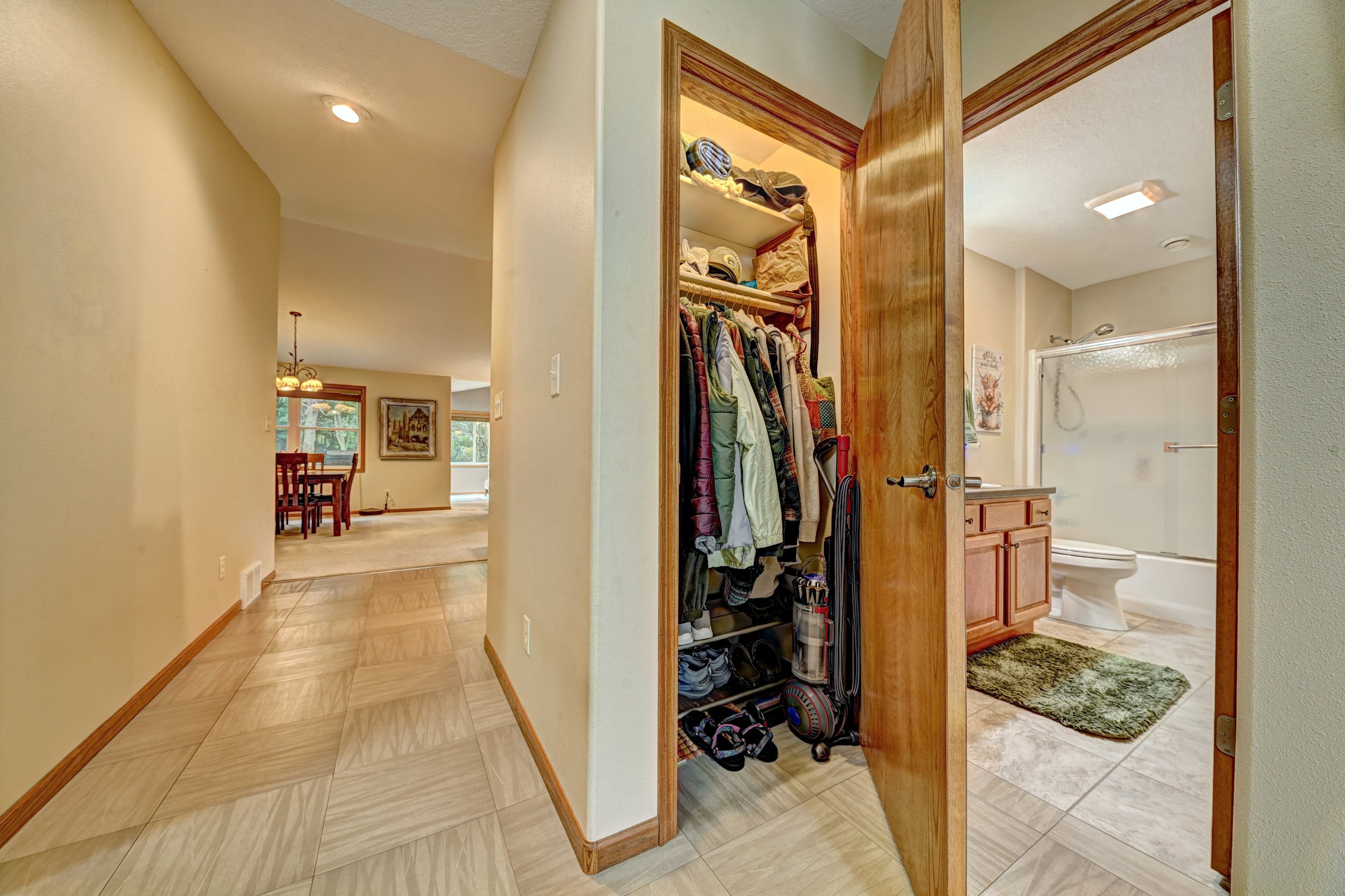 A view of a home's interior, showing a hallway leading to a dining area, with an open closet filled with clothes and items, and part of a bathroom with a shower and toilet visible through an open doorway.
