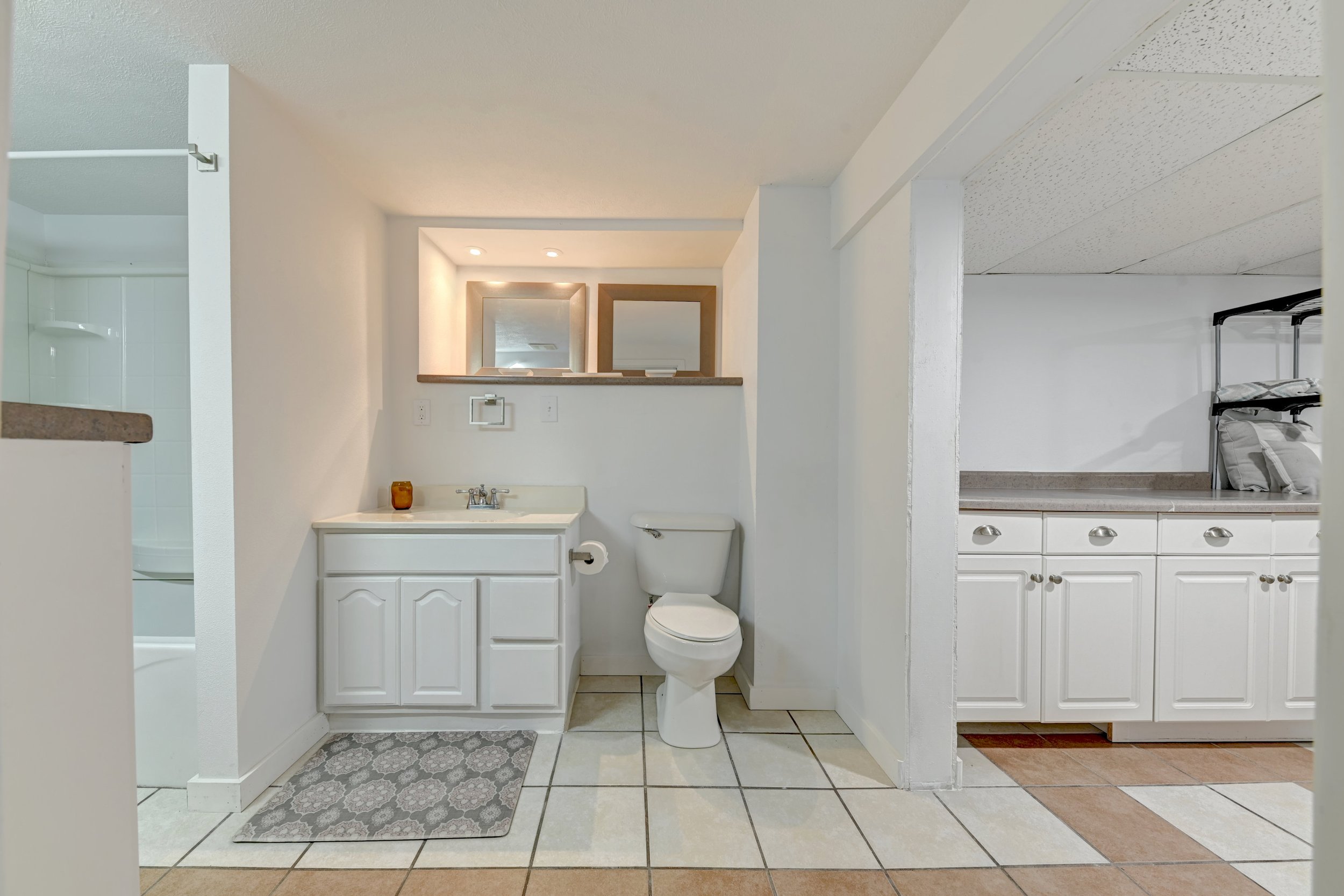 A small bathroom with a white vanity, a toilet, and a mirror above the sink. To the right is part of a kitchenette with a countertop, cabinets, and some stacked bedding. To the left, there is a partial view of a shower. The floor has white and beige 