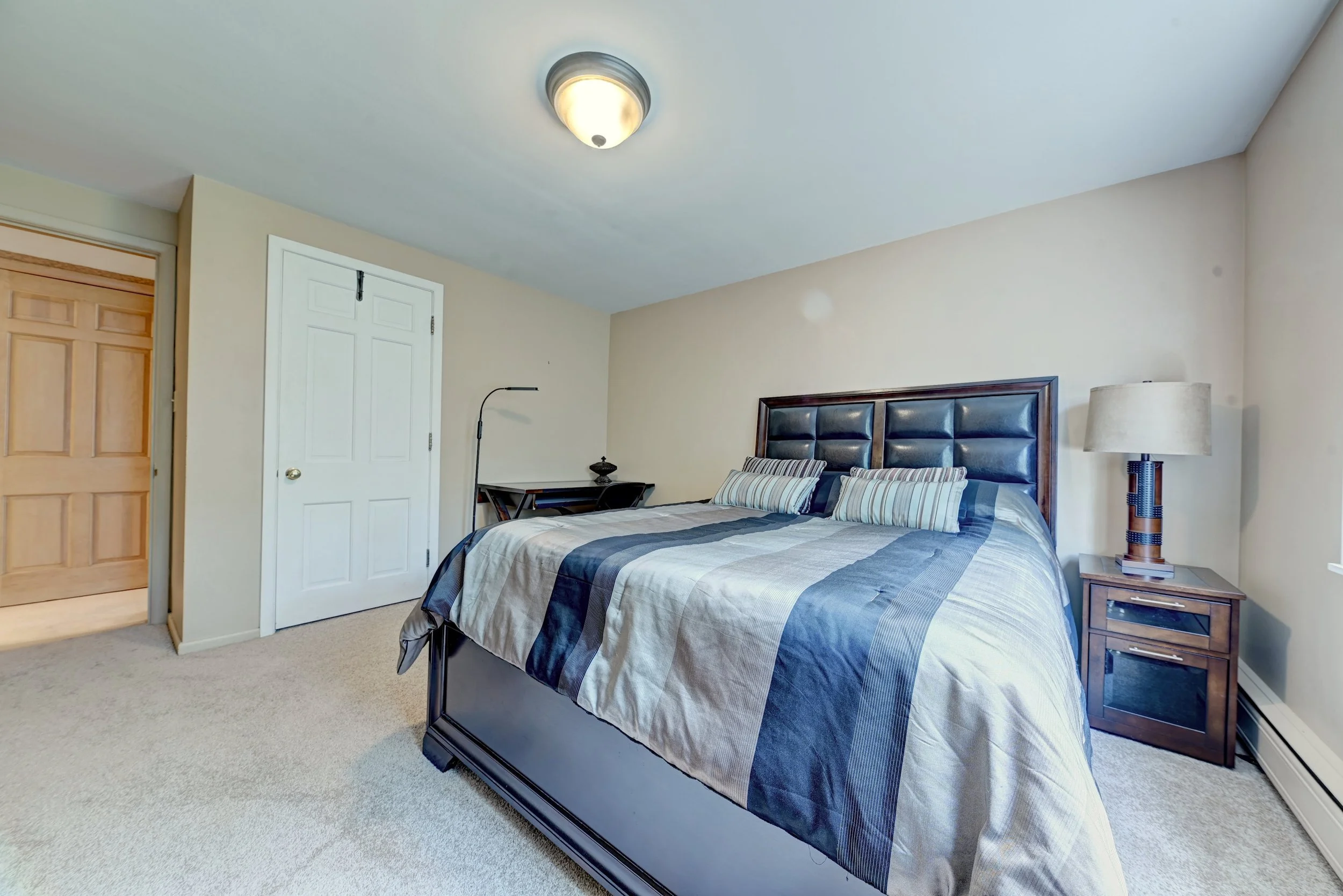 A bedroom with a bed, nightstand, and lamp. The room has beige walls, a wooden door, and a white closet door.