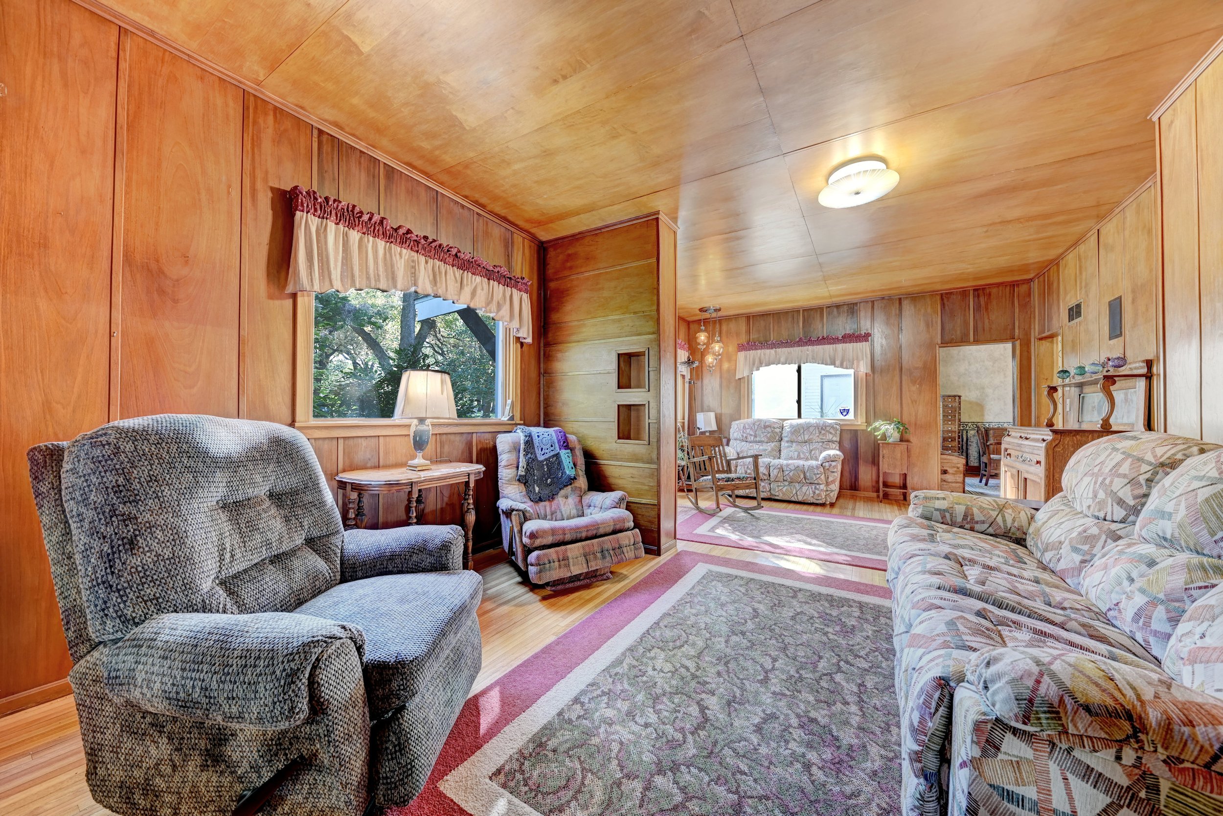 Living room with wood-paneled walls, multiple chairs, a sofa with a patterned slipcover, and a large window with curtains overlooking trees.