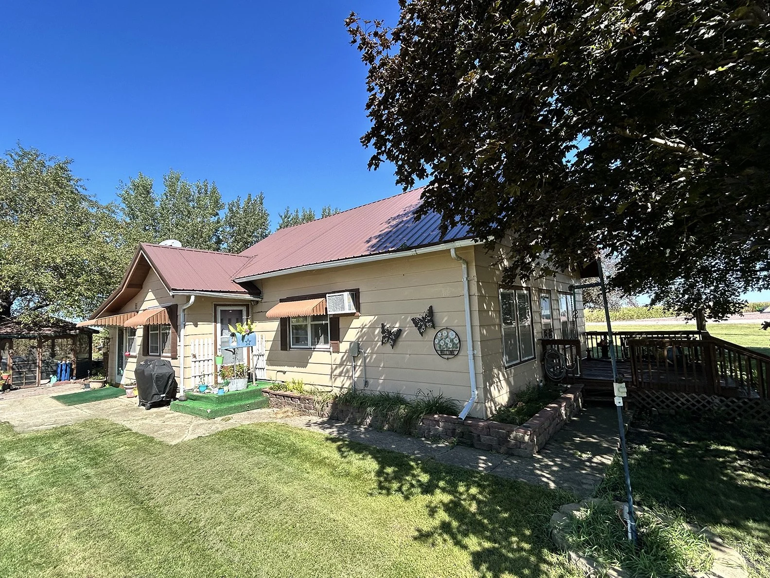 A small house with a red metal roof, beige siding, and several windows with brown shutters and awnings. The yard has a lawn, a small garden bed, and decorative butterfly and floral wall hangings. There is a wooden deck on the right with a black raili