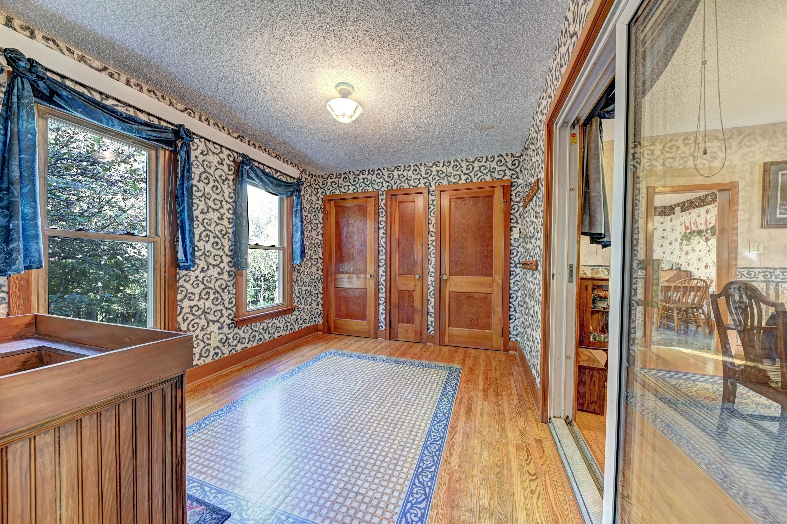 A room with hardwood floors, patterned wallpaper, and a ceiling light. There are windows with blue curtains, wooden doors, and a glass sliding door leading to a dining area with a table and chairs.