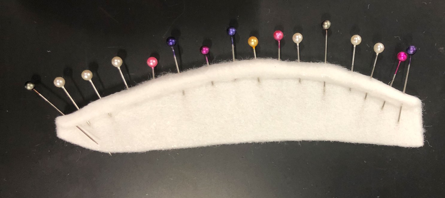 A piece of Fosshape, a white felted looking fabric, with one edge rolled in on itself. The rolled portion is held in place with sewing pins.