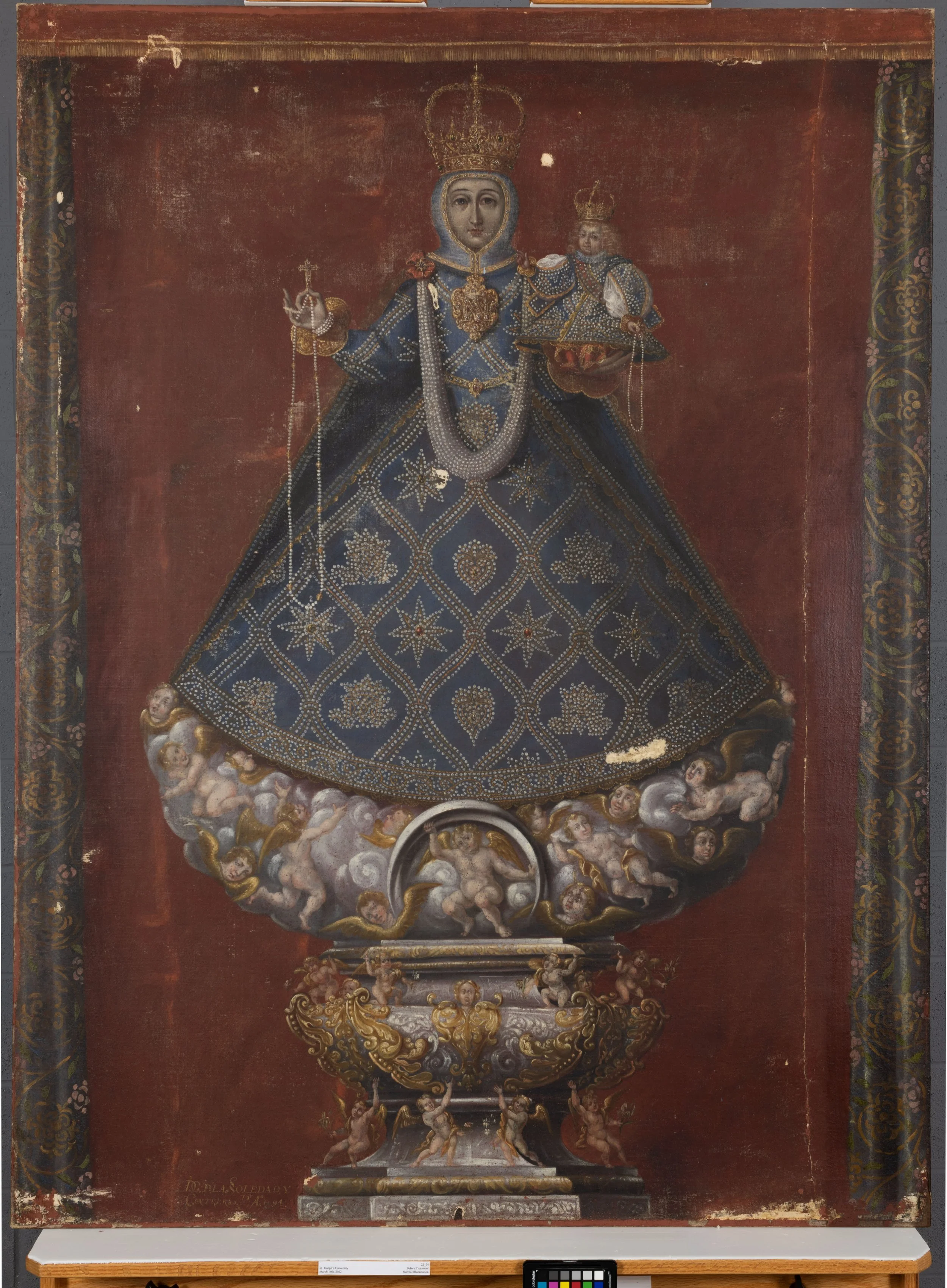 Painting of the Madonna holding the Baby Jesus while standing on a white pedestal with a dark red background. Both are wearing highly decorated and patterned blue gowns and gold crowns. Photo is before treatment showing damage and paint loss.
