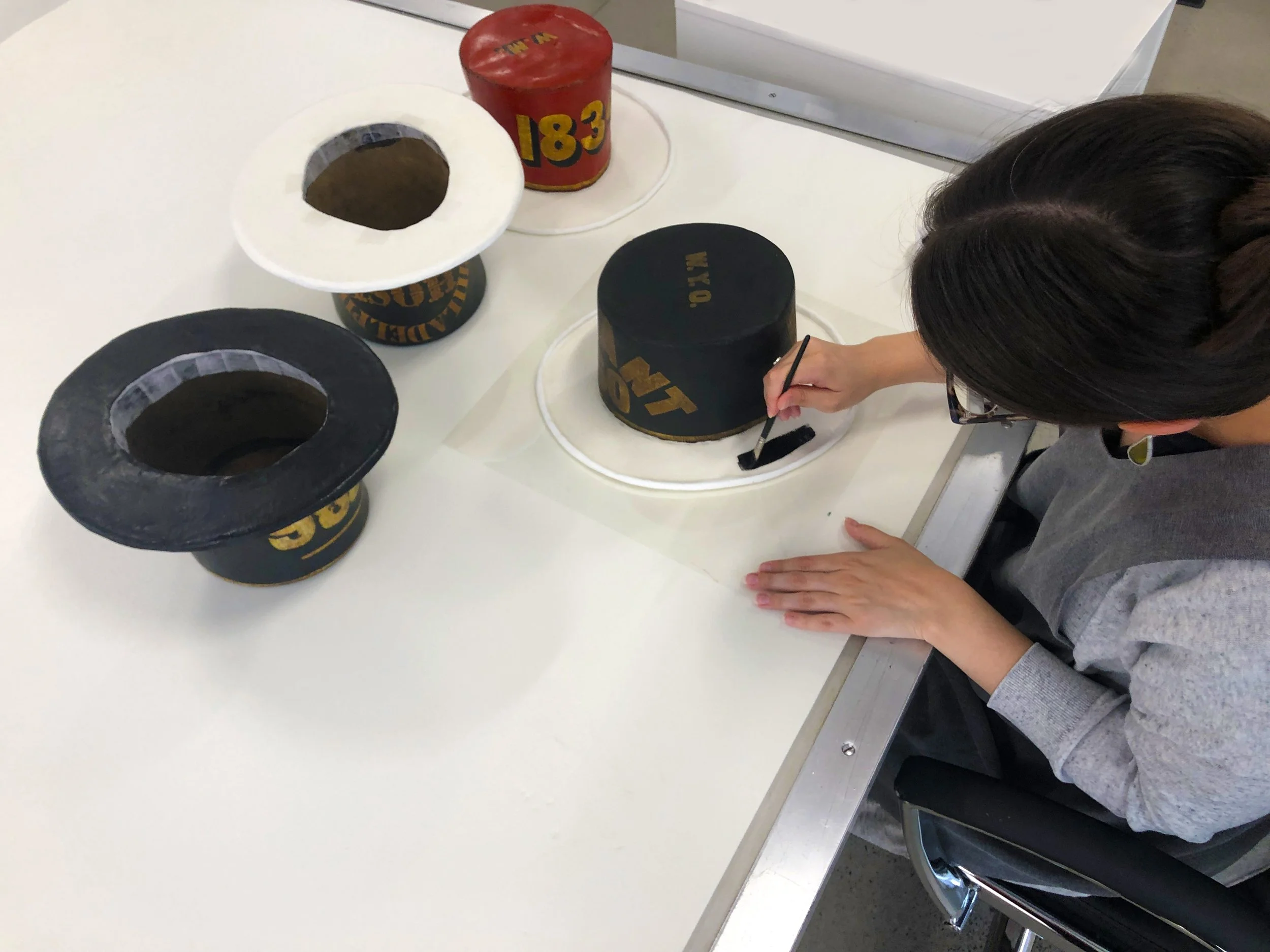 A conservator sitting at a table painting the white brim she created for a parade hat as part of treatment the same color as the original stove pipe portion of the hat. Three other hats also undergoing treatment are on the table.