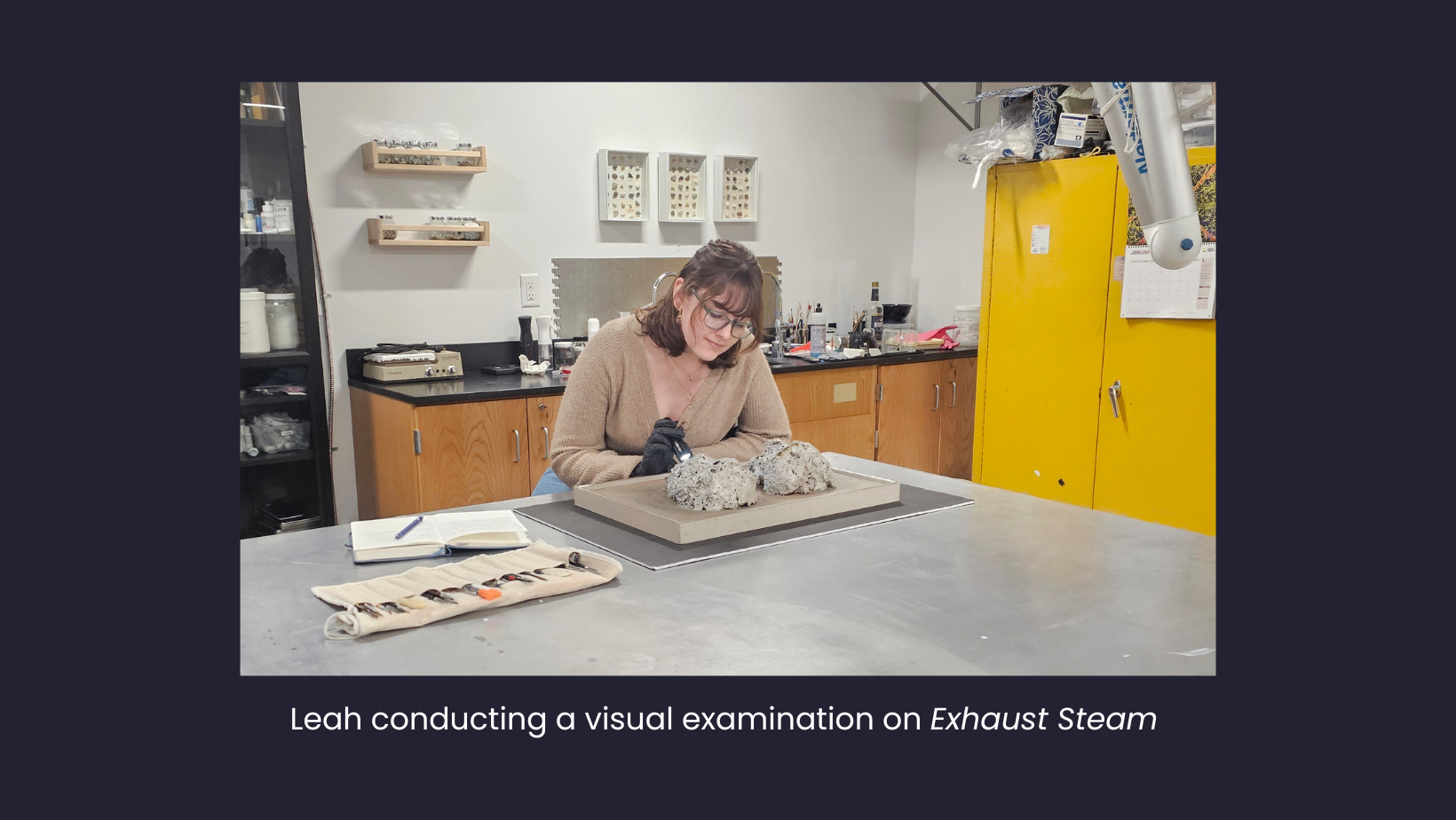 Leah sitting at an aluminum topped table using a flashlight to visually inspect a painting on the table. A set of conservation tools and an open notebook with a pencil are on the table next to the painting.