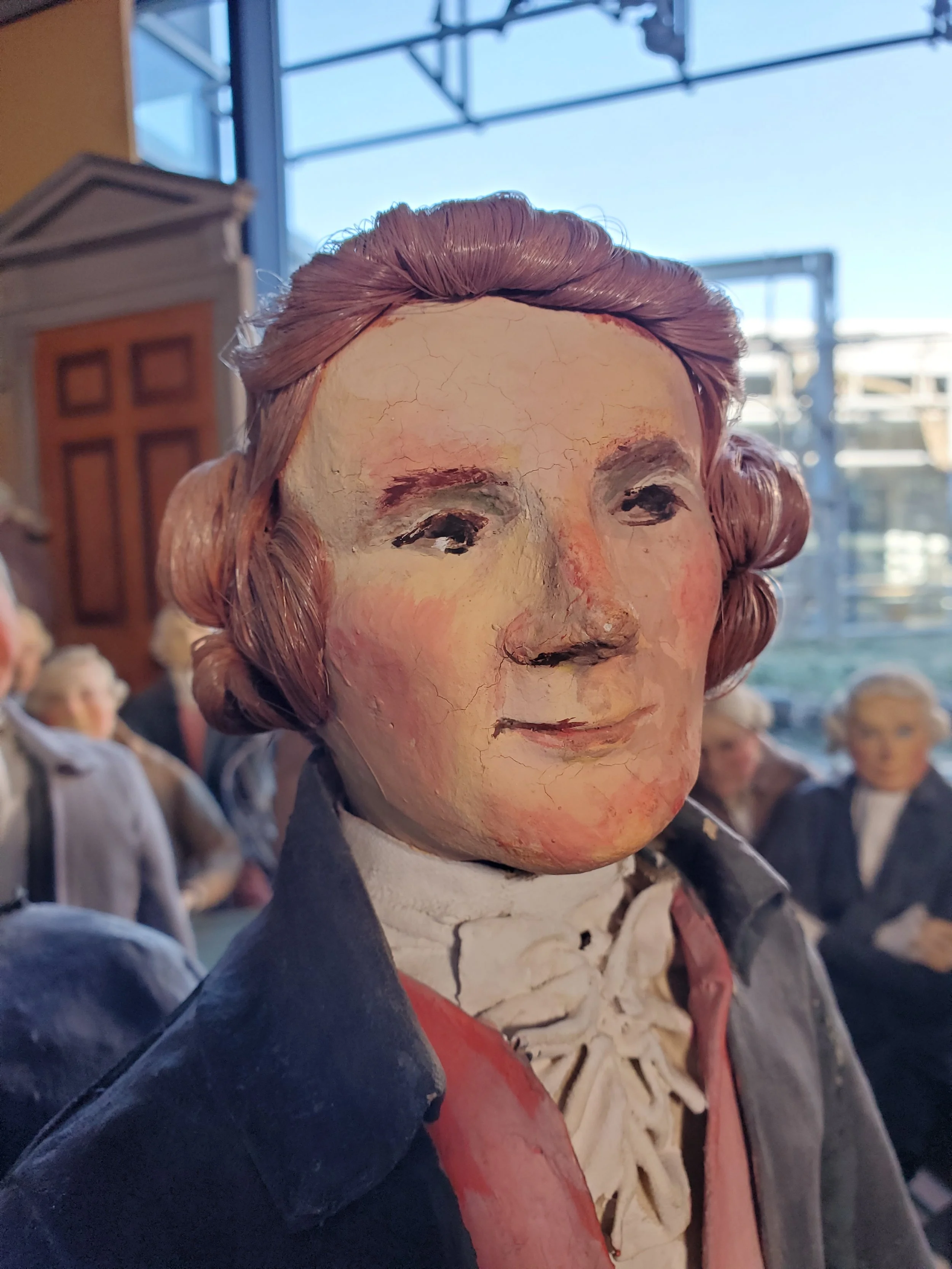 Detail photo of the Thomas Jefferson figure in the diorama from the chest up. The figure is wearing a blue coat, red vest, and white undershirt.