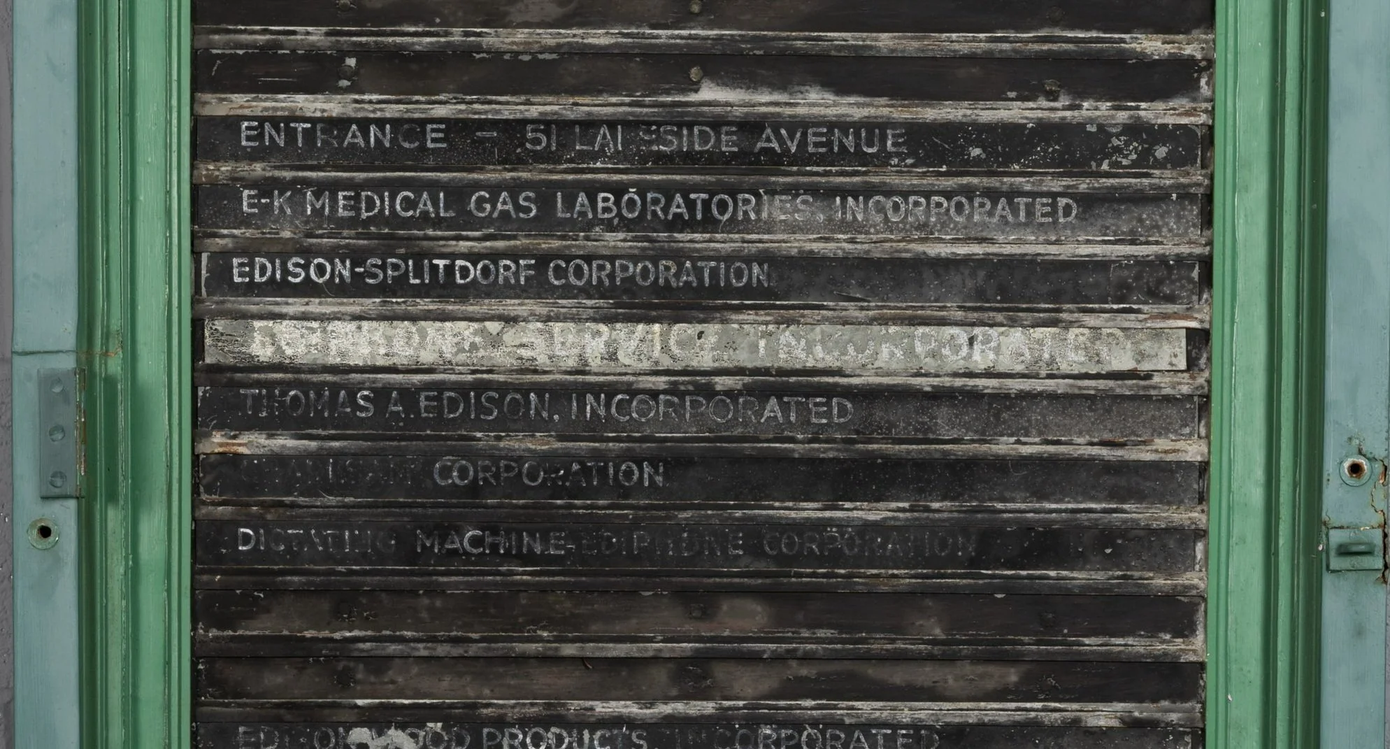 A historic directory sign with the names of companies in white paint on a black background. Both the white and black paint have faded due to long term light exposure, making the words hard to read.