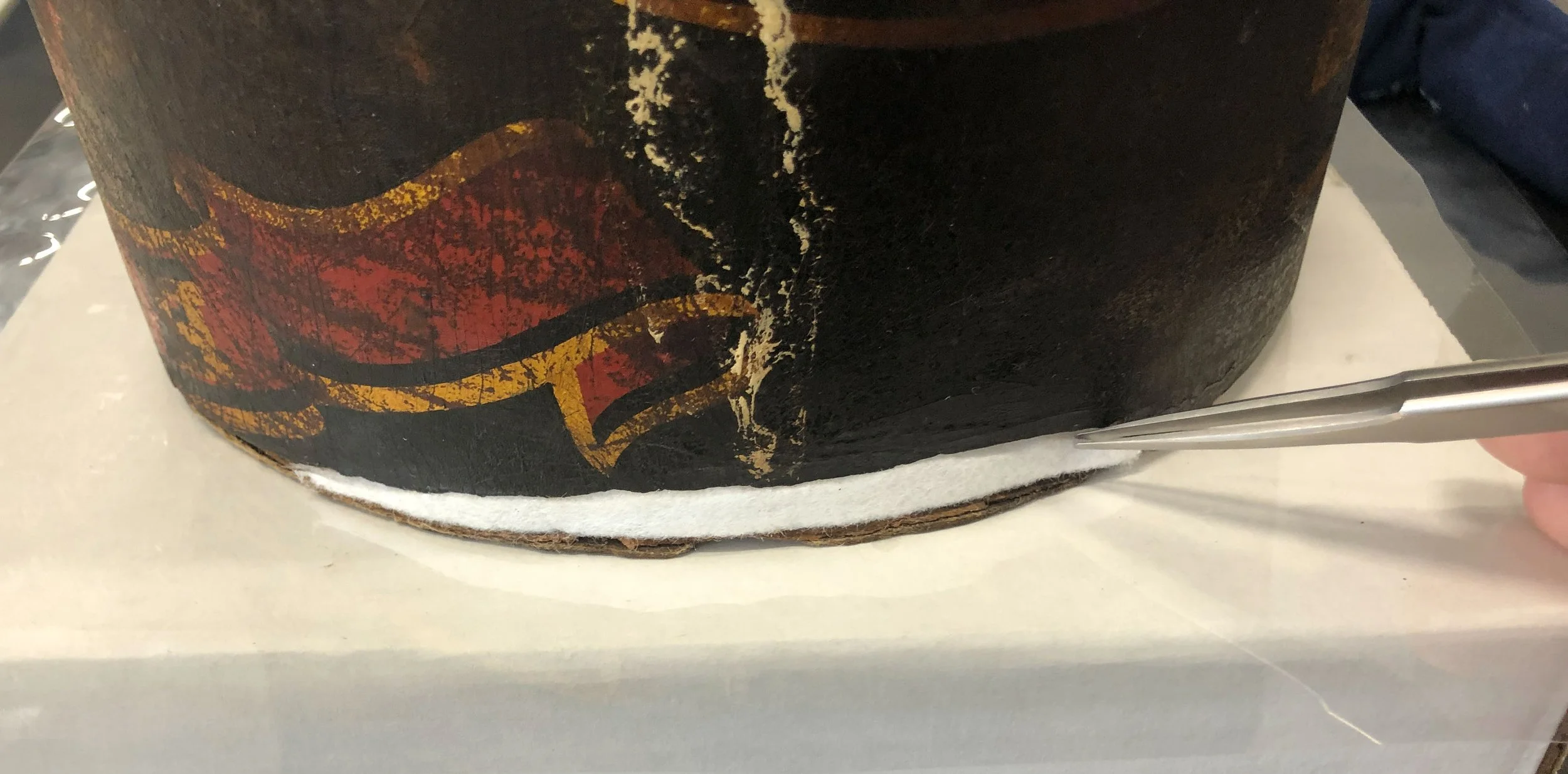 A piece of white felted fabric is placed into an area of loss on a black damaged and worn hat by a pair of tweezers to repair that area as part of the conservation treatment