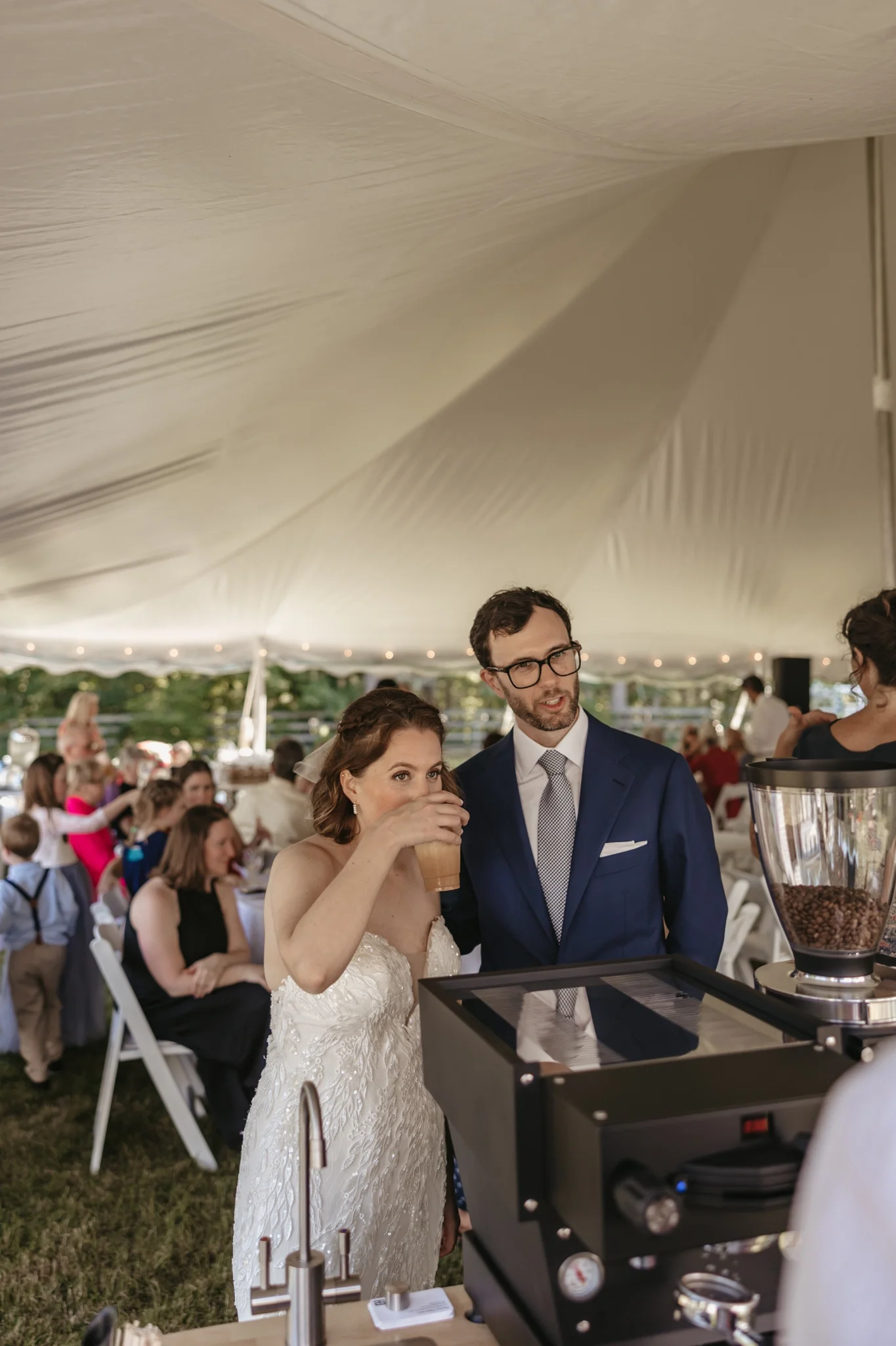 Bride and groom at wedding reception, getting coffee from a bar under a large tent, guests seated at tables in the background.