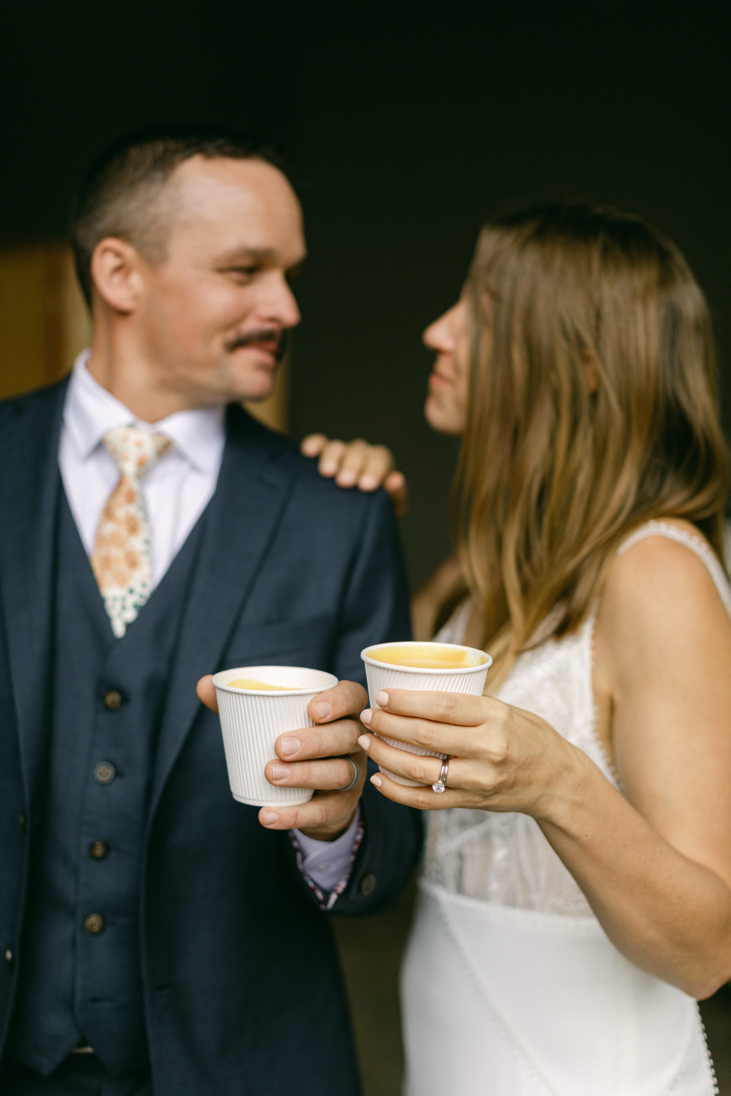 A man and woman in formal attire holding cups of coffee, standing close and looking at each other.