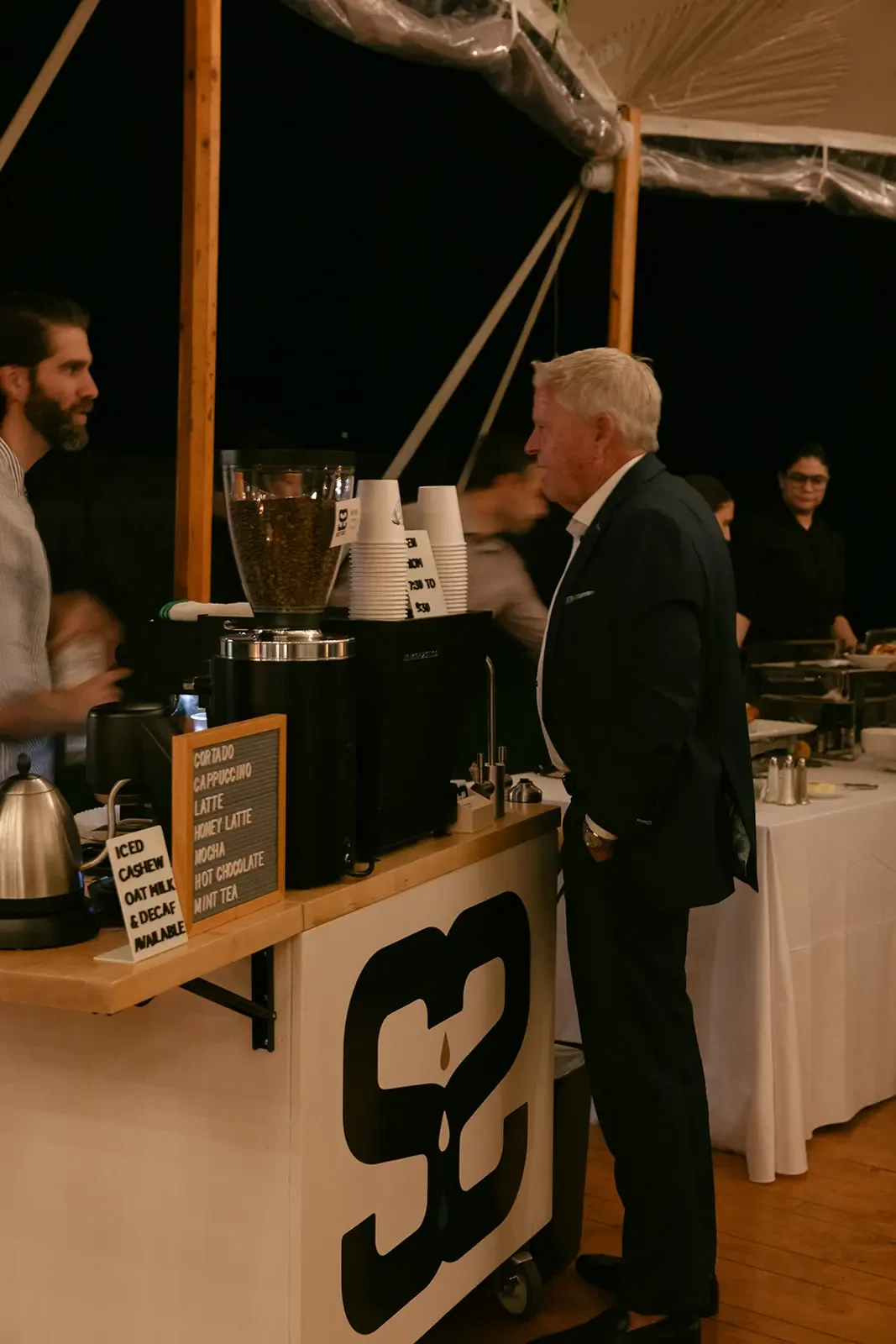 An elderly man in a black suit ordering coffee at a nighttime outdoor coffee stand. The counter features a large coffee bean container and a sign with drink options.