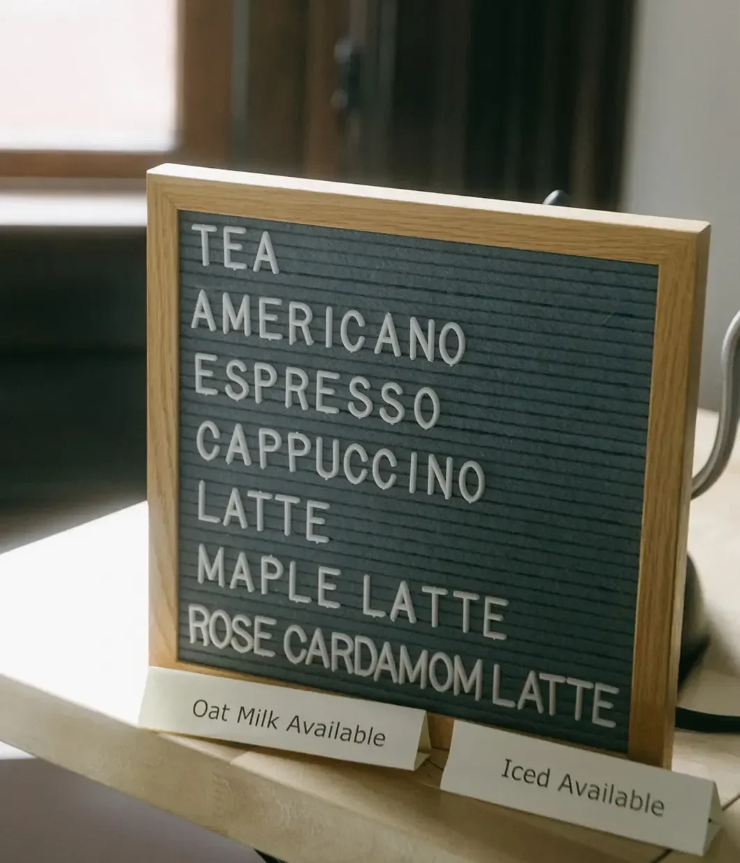 Menu board with drink options including tea, americano, espresso, cappuccino, latte, maple latte, and rose cardamom latte, with notes indicating oat milk and iced options available.