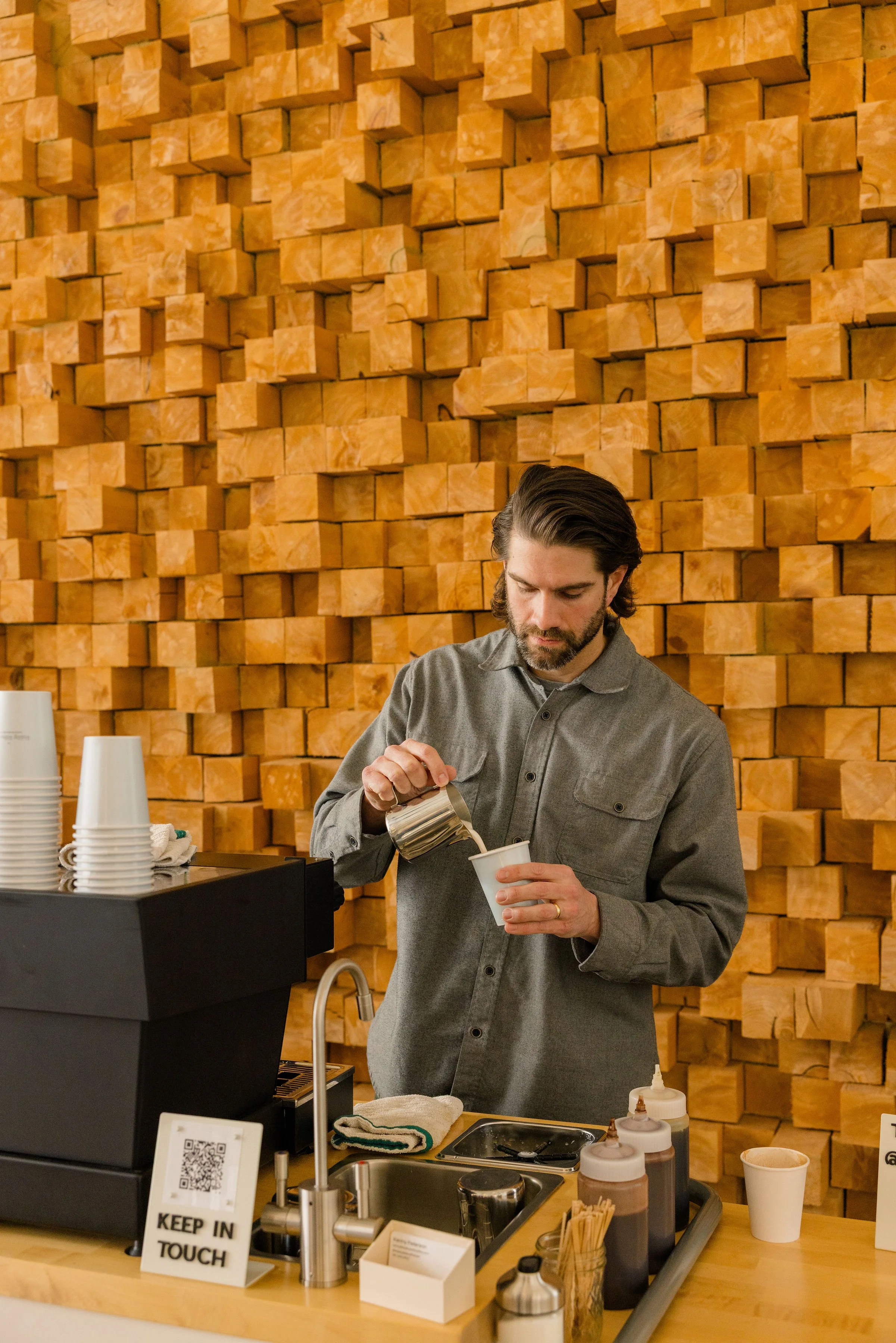 A man with dark hair and a beard, wearing a gray shirt, prepares a hot beverage at a coffee shop counter. Behind him is a textured wall made of stacked, uneven wooden blocks.