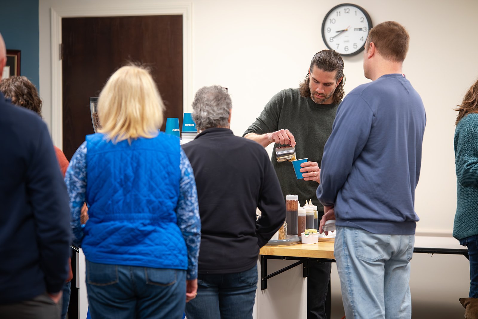 People lined up at a coffee station in a room, with a man serving coffee to a customer, and old-style clock on the wall showing about 12:15.