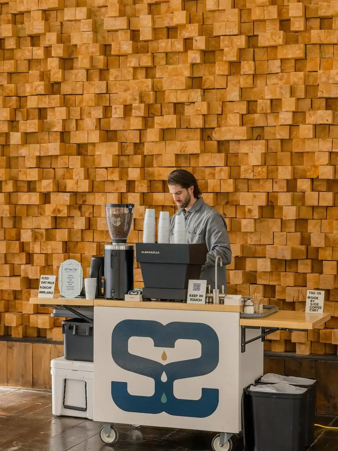 A coffee stand with a man preparing drinks in front of a brick wall, on a cart with a large coffee logo, coffee cups, and a blender.