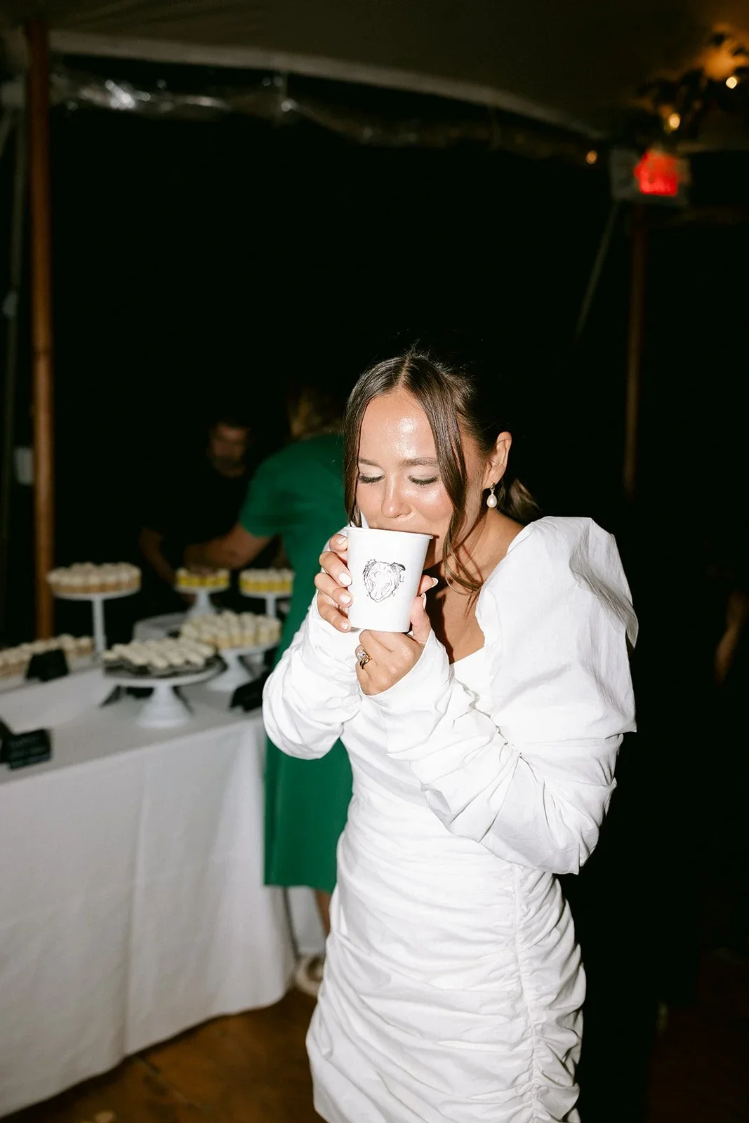 A woman in a white dress enjoying a beverage at a celebration or party, with a dessert table in the background.