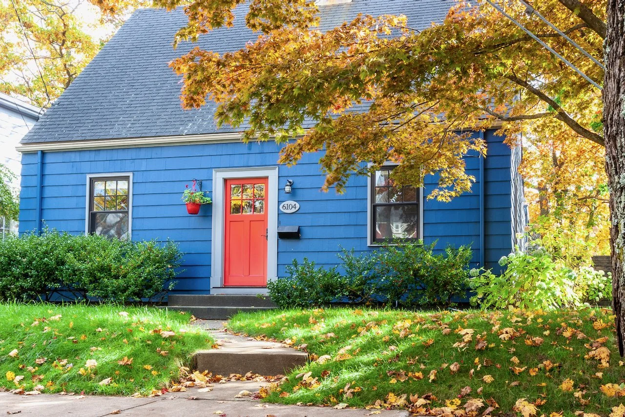 A blue house with a red front door, surrounded by green bushes and trees with autumn leaves, on a sunny day.