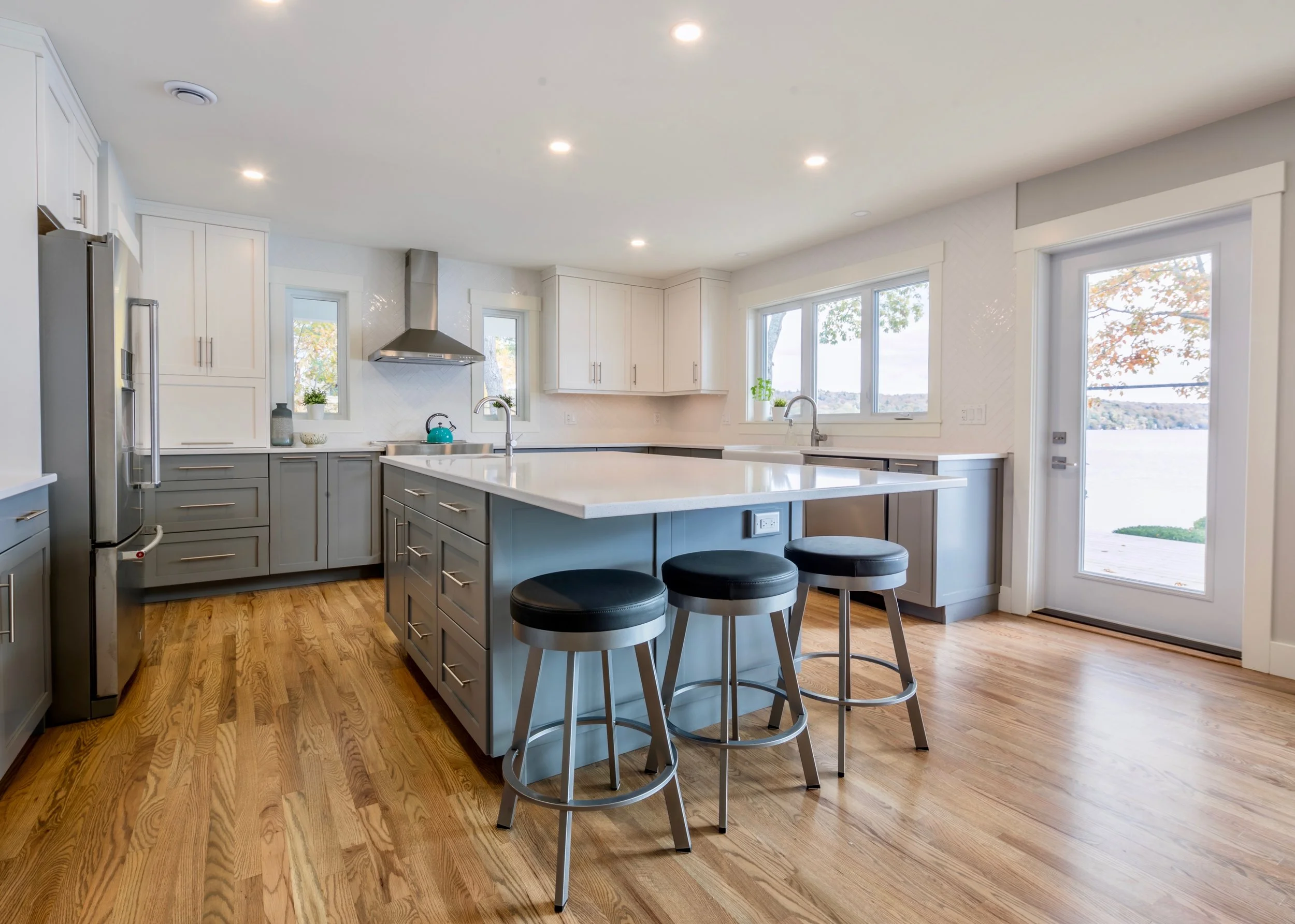 Modern kitchen with white and gray cabinetry, hardwood floors, and a large window view of trees and water outside.