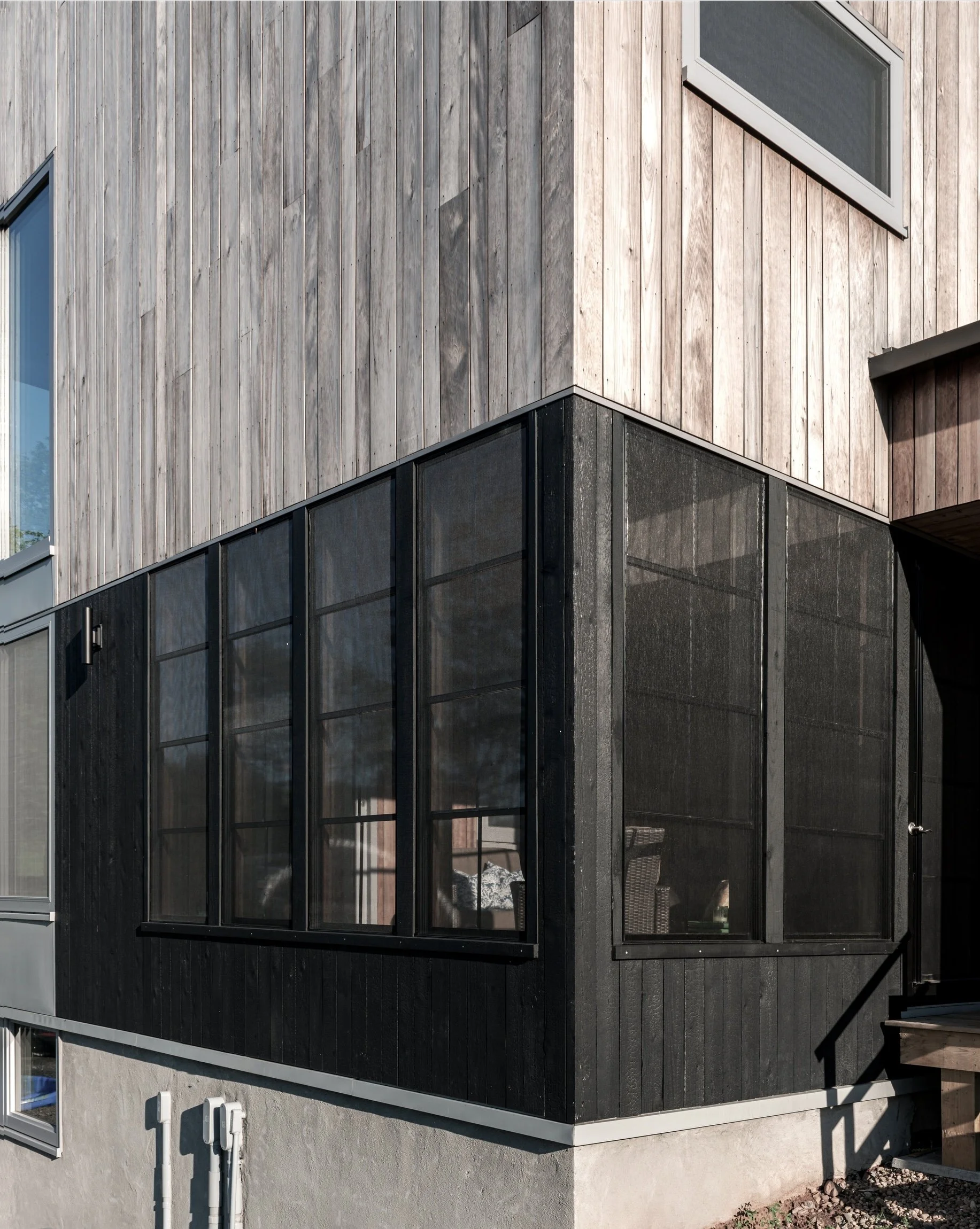 The building's exterior features a modern design with a combination of materials. The upper section is clad in vertical wooden planks with a weathered finish, while the lower section consists of large, dark-framed windows. There is a small overhanging roof section, and a staircase with a metal railing is partially visible on the right side of the image.