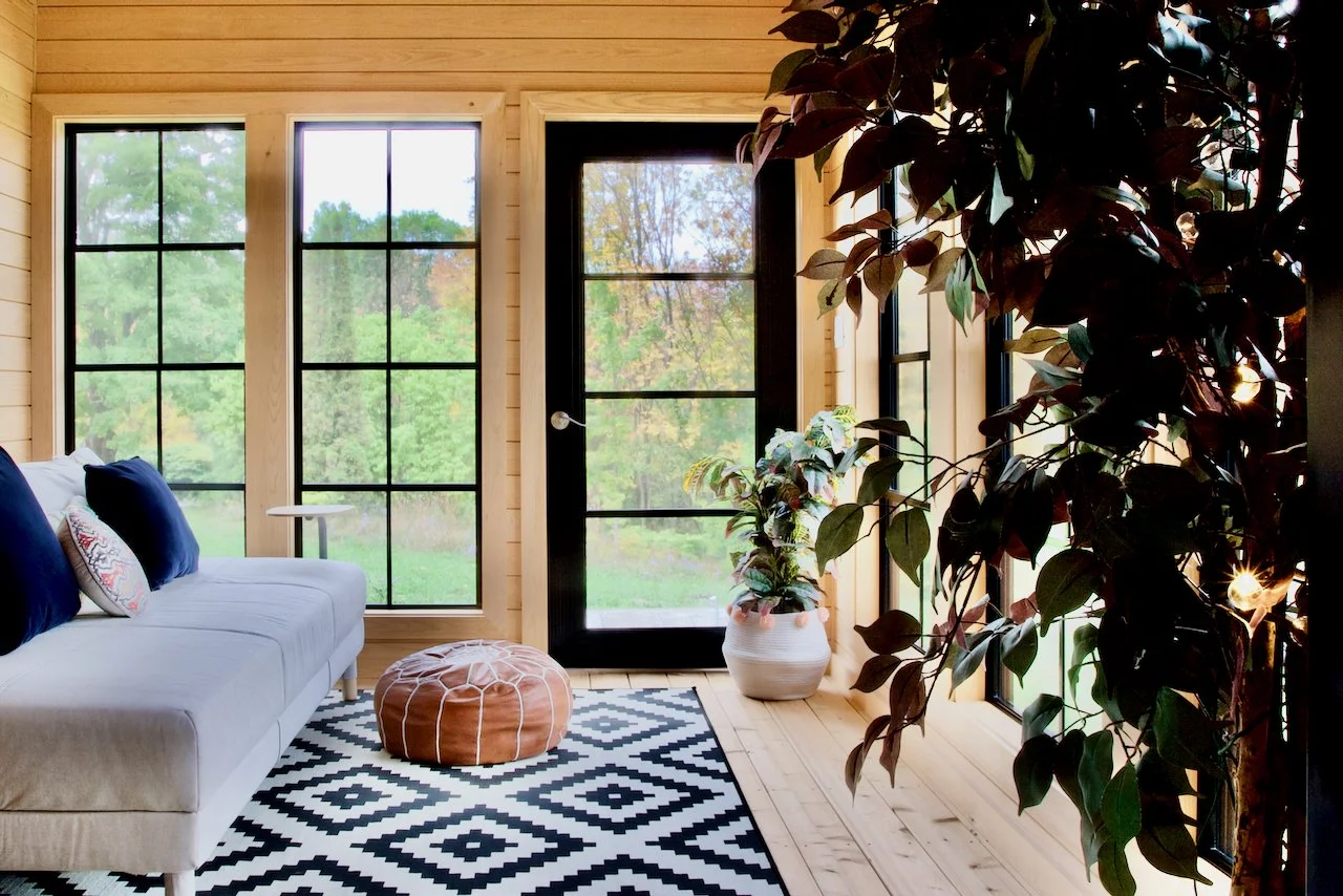 A cozy living room with large windows showing greenery outside, a white couch with dark blue pillows, a leather pouf, a black and white patterned rug, and potted plants inside a wooden-paneled space.
