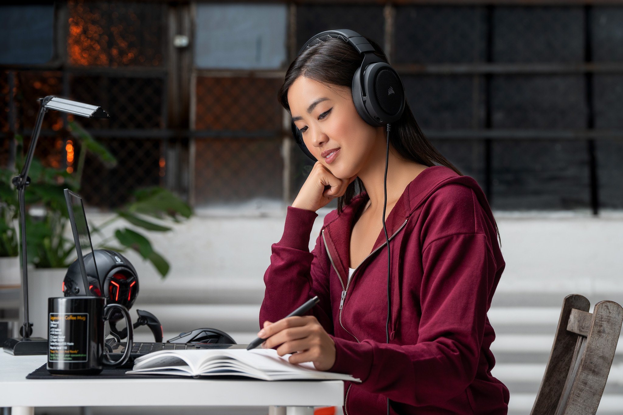 Woman with long dark hair wearing headphones, a maroon zip-up hoodie, sitting at a desk, writing in a notebook, with a computer, headphones, mug, and plants on the desk.