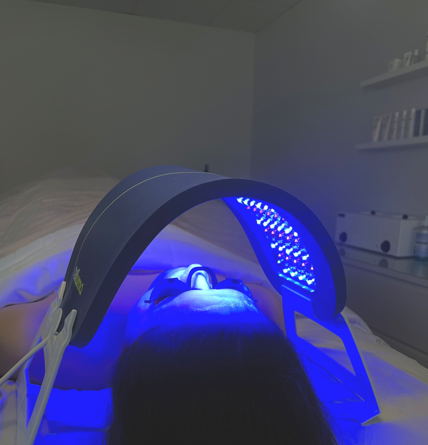 Person undergoing LED light therapy treatment with their face illuminated by a blue LED mask while lying on a bed in a medical or spa room.