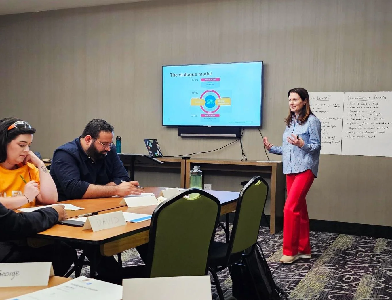 A woman giving a presentation to a small group in a conference room with a whiteboard and a large screen displaying a diagram.