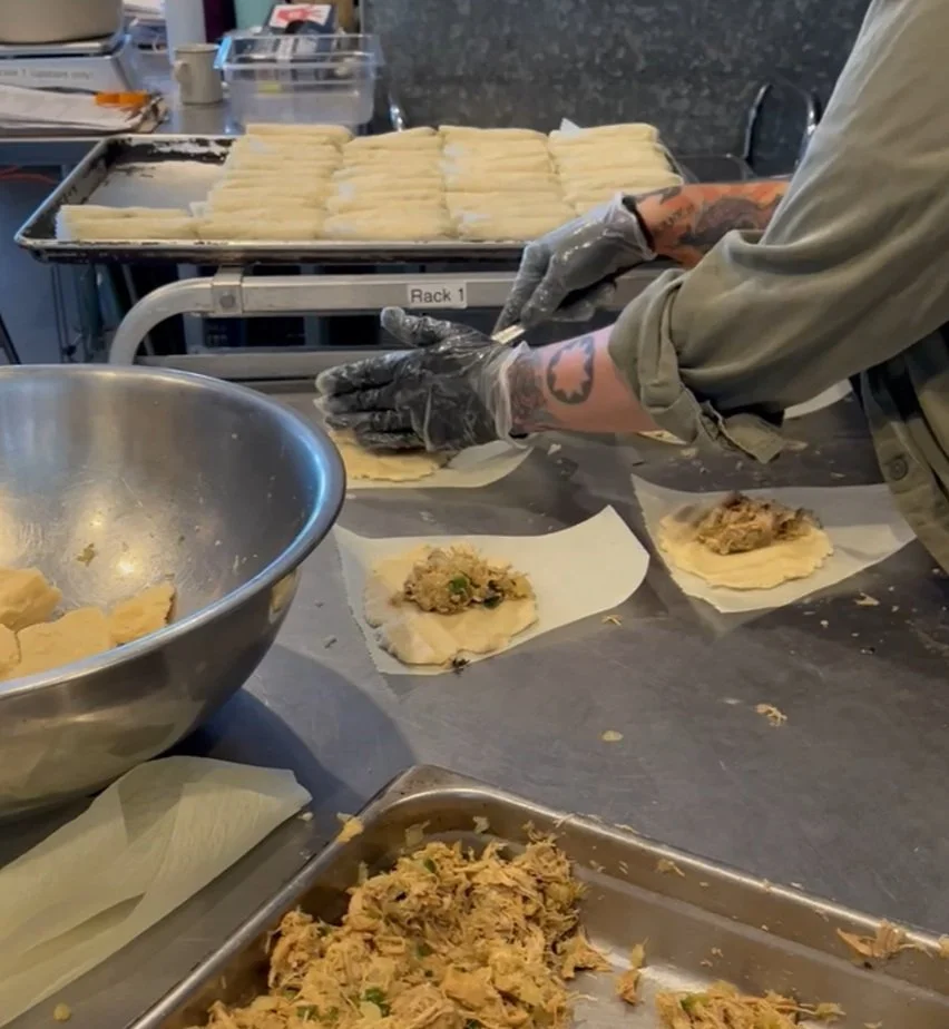 Person preparing dough with meat filling on a counter, wearing gloves, with a tray of prepared dough in the background.