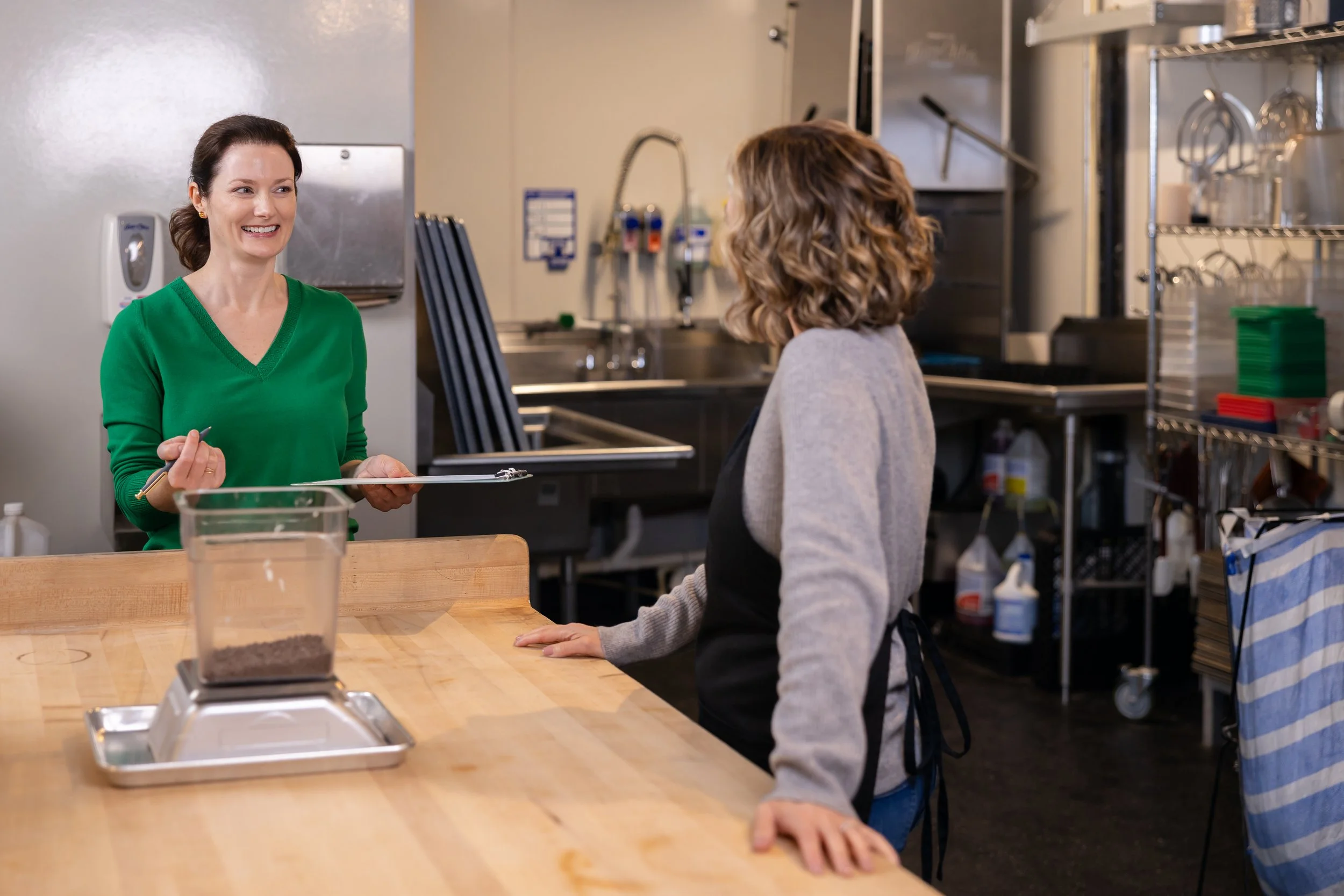 Two women in a commercial kitchen engaged in a conversation. One woman, wearing a green sweater, is smiling and holding a pair of tongs, while the other woman, with curly hair and a gray sweater, stands at a wooden counter with her hand resting on it.