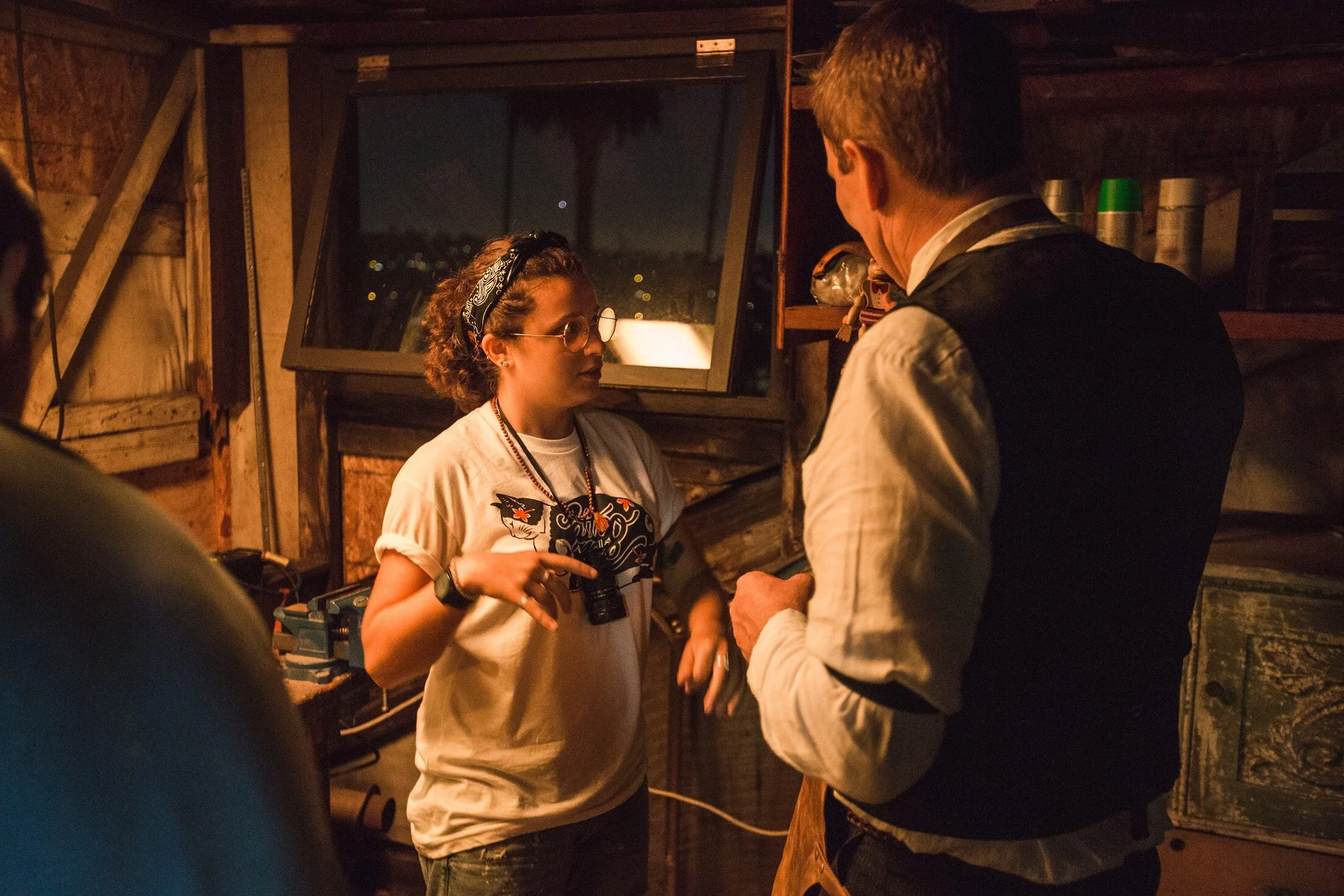 Two people having a conversation inside a wooden shed at night, with a window showing a dark sky and city lights.