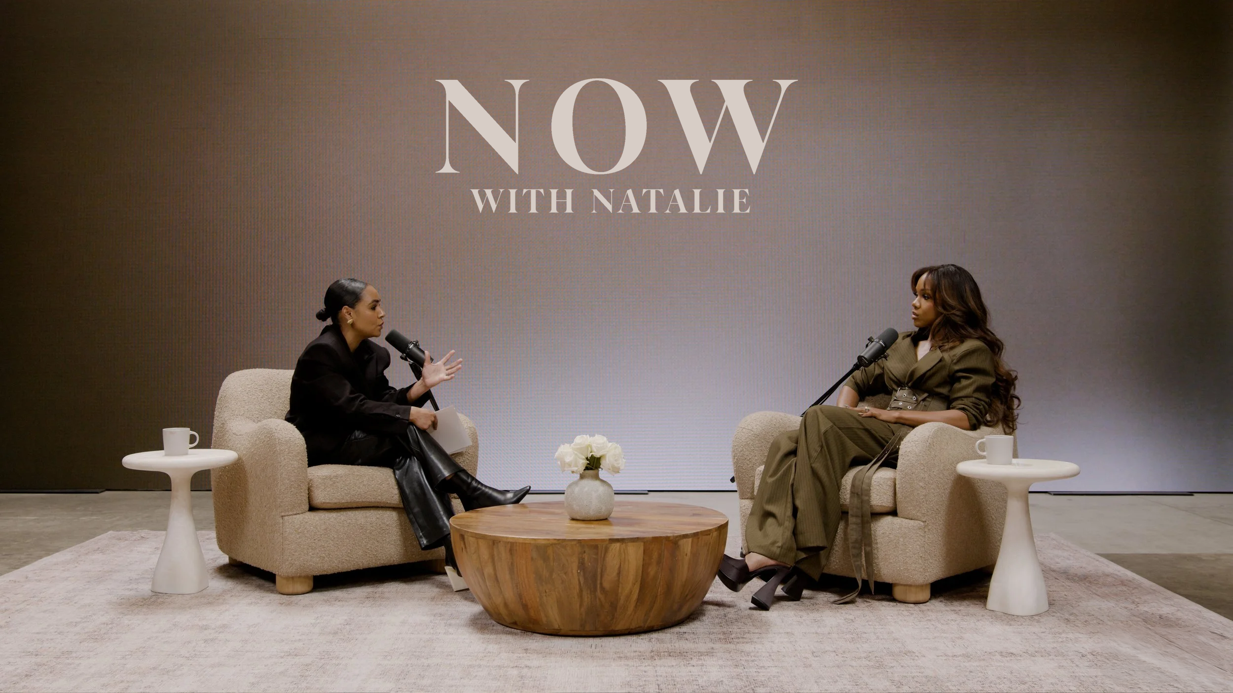 Two women sit in beige armchairs, having a conversation in a TV or podcast interview setup. They are separated by a wooden round coffee table with a white vase of flowers. Each woman has a microphone and a white coffee mug on a small white side table next to her. Behind them is a large screen displaying the words 'NOW WITH NATALIE'.