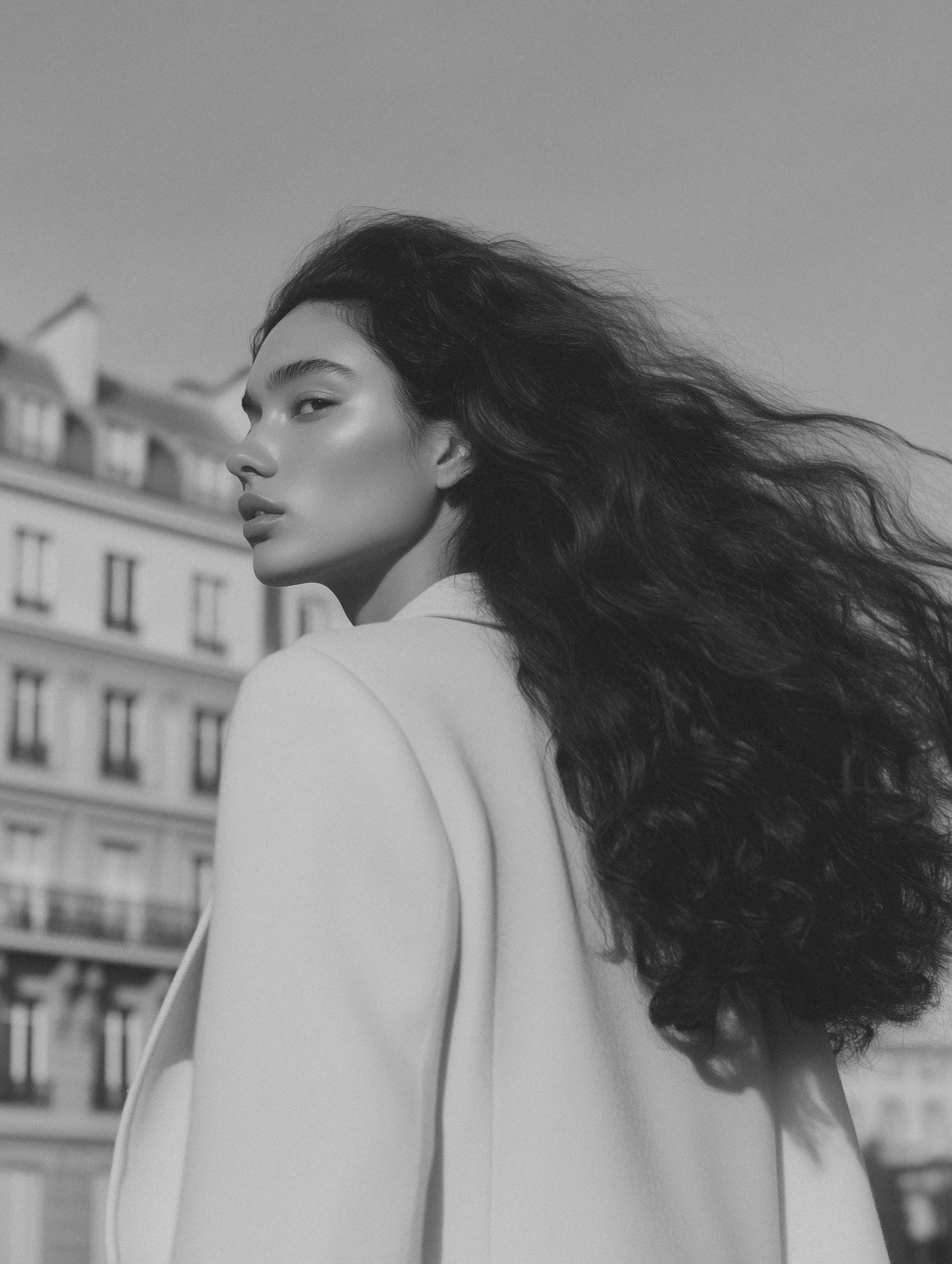 Black and white photo of a woman with long, wavy hair in an outdoor urban setting, looking over her shoulder.