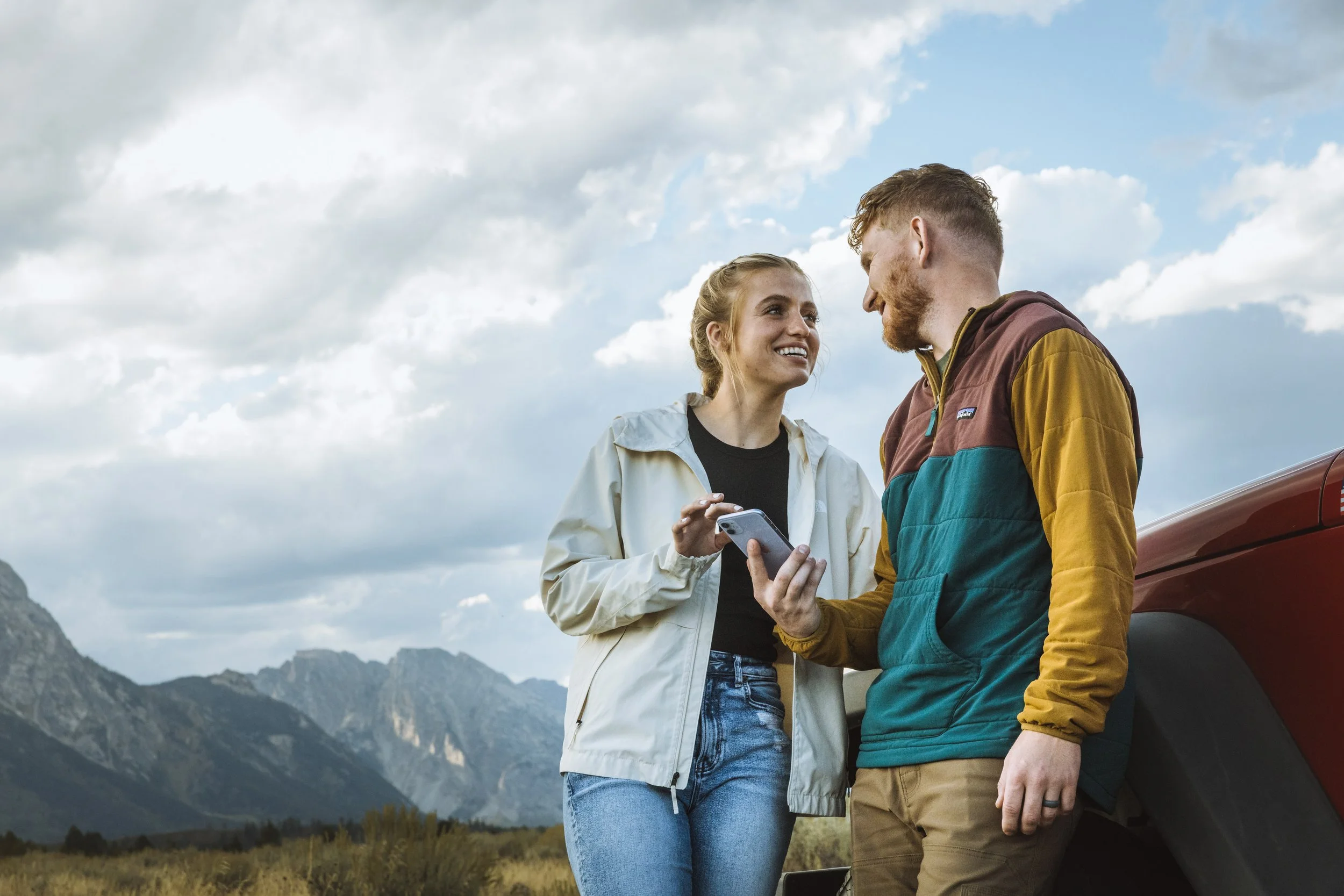 A young woman and man standing outdoors near a red vehicle, smiling and looking at each other, with mountains and a cloudy sky in the background.