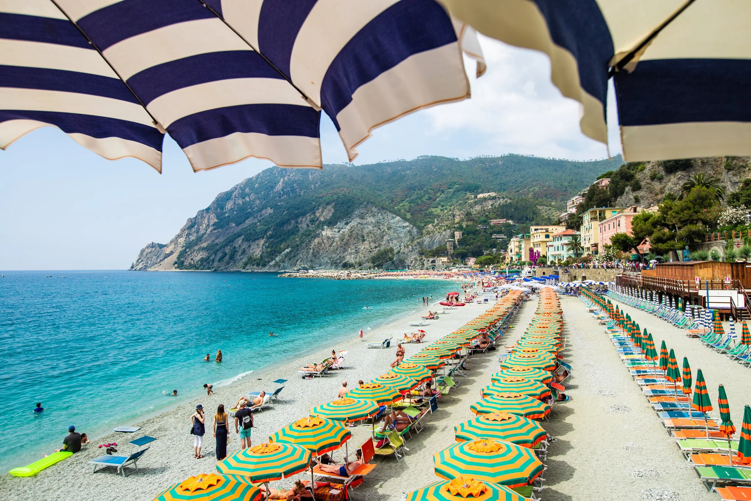 Beach scene with colorful umbrellas, people swimming and relaxing by the water, hillside with houses in the background.