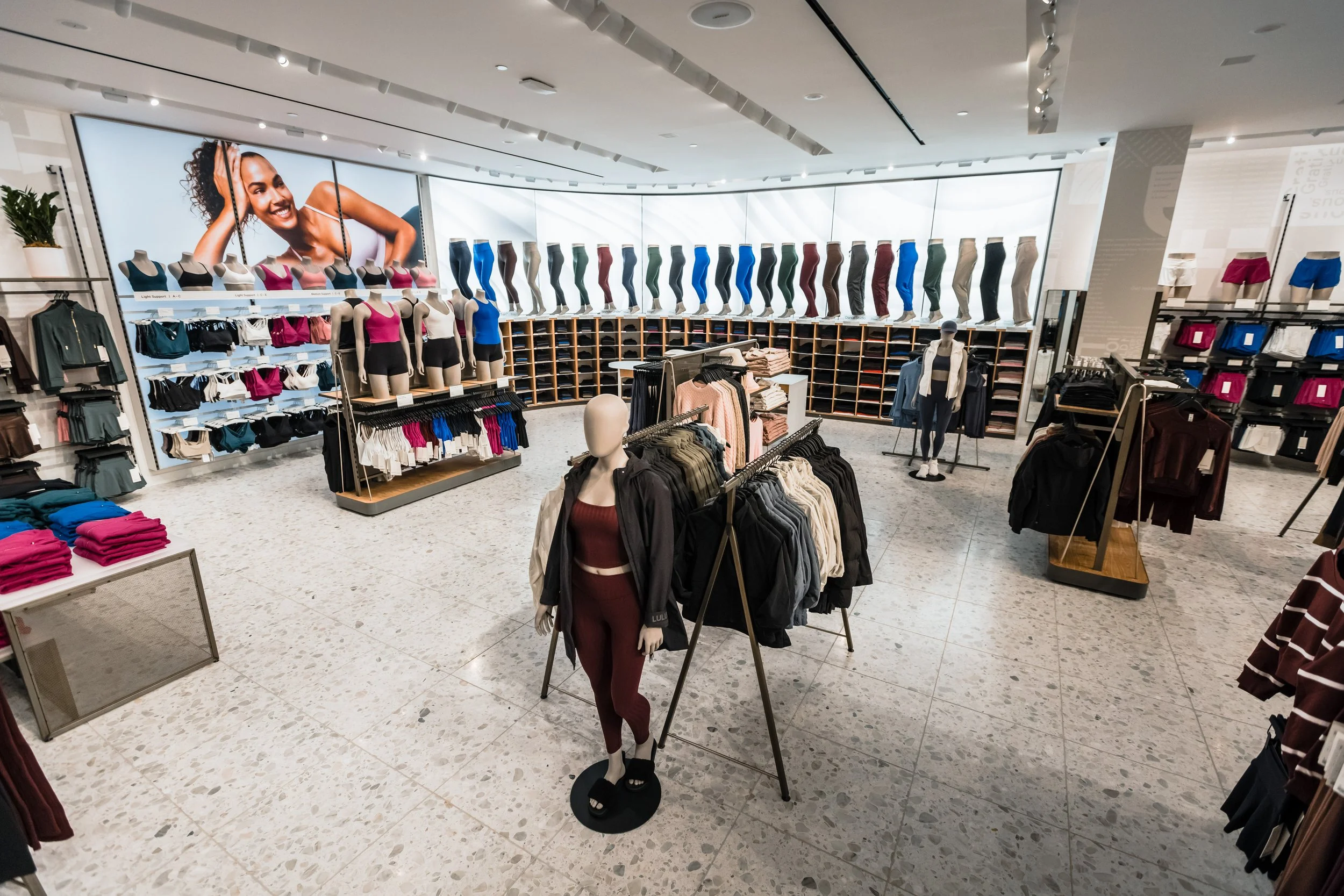 A clothing store displaying various athletic apparel, including mannequins dressed in workout outfits, shelves with folded clothes, and a large digital screen showing a smiling woman in activewear.