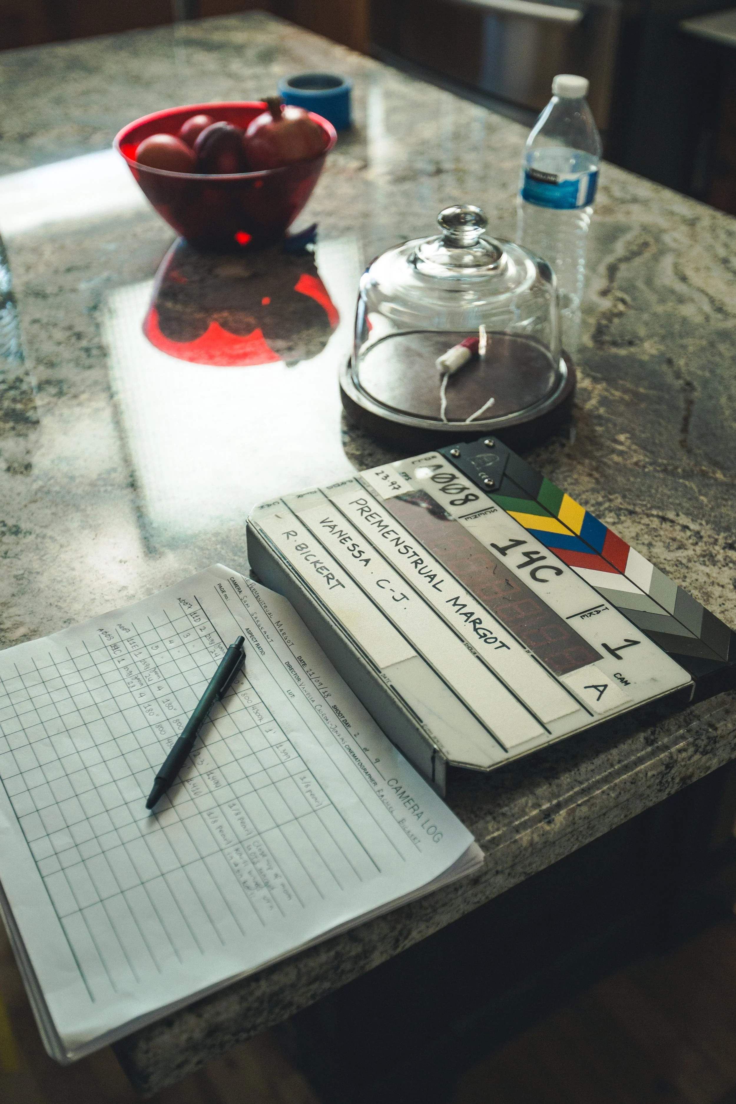 Clapperboard, script, and pen on granite countertop with a bowl of eggs, plastic water bottle, and glass cake dome in background.