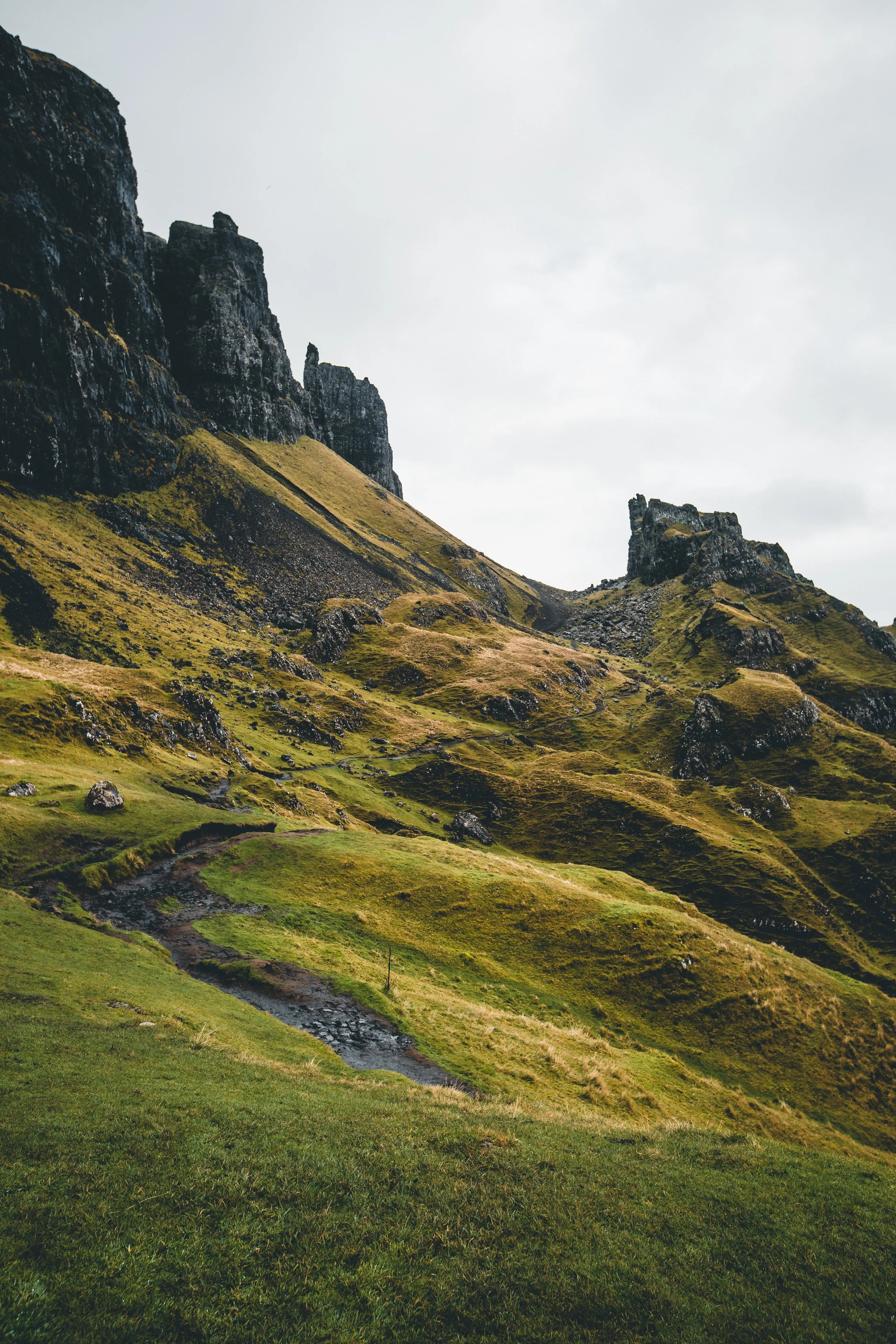 Scenic view of a grassy hillside with patches of rocks and a steep mountain in the background under a cloudy sky.