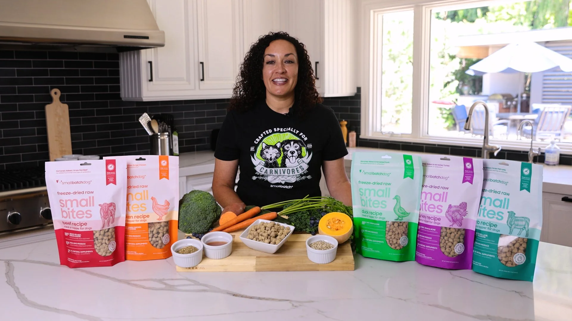 A woman standing in a kitchen with various pet food ingredients and bags of Small Bites freeze-dried raw dog food in front of her.