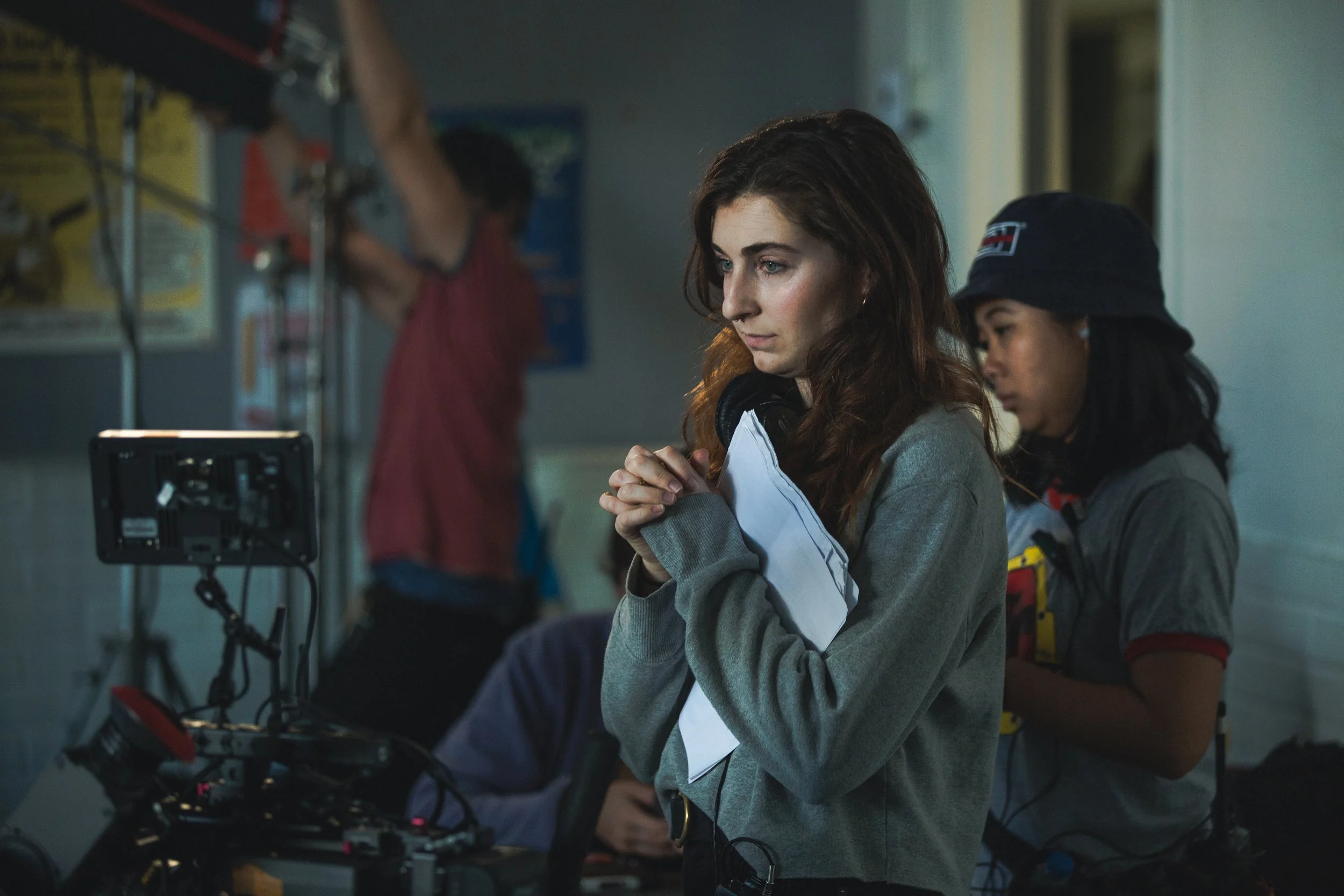 A young woman with brown hair sits with her hands clasped and eyes closed, wearing headphones around her neck, in a filming or production studio. In the background, two other women are working with filming equipment.