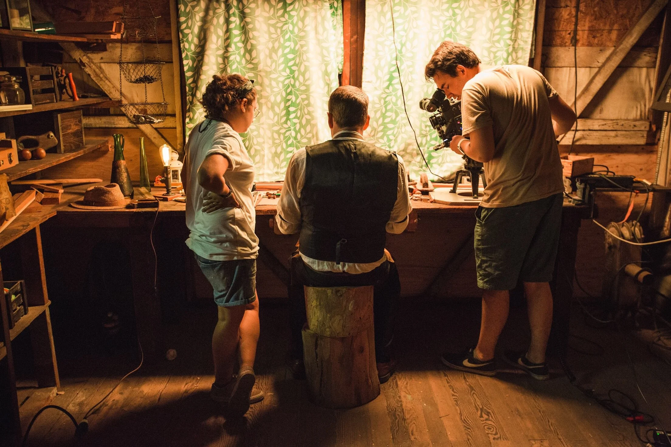 Four people working on a film set in a rustic wooden workshop with green patterned curtains and shelves of tools and supplies. One person is seated, filming with a camera mounted on a tripod, while two others are standing nearby, assisting or observi