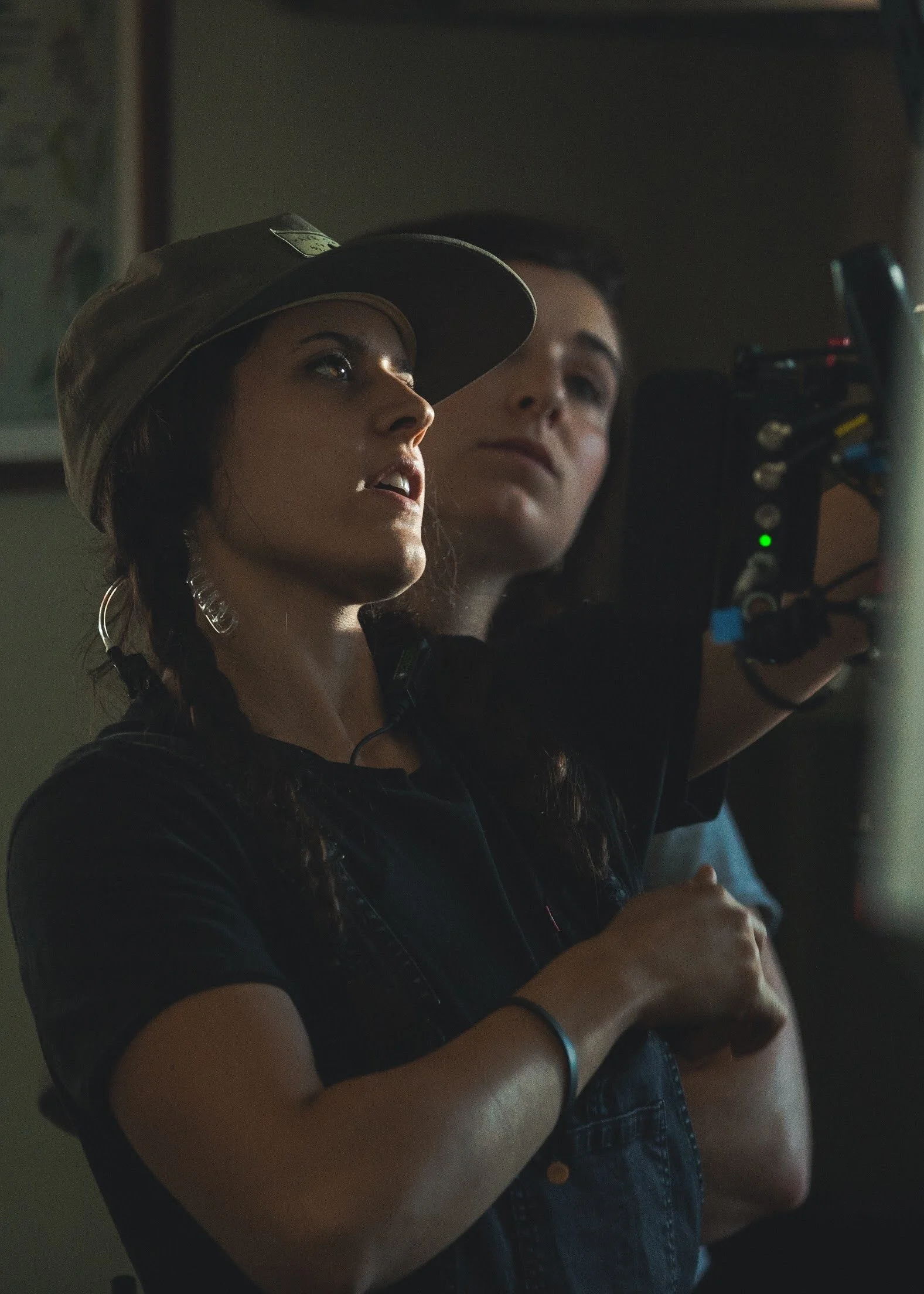Two women are filming a scene with a camera; one woman is wearing a hat and has braided hair, the other woman is standing behind her, observing.