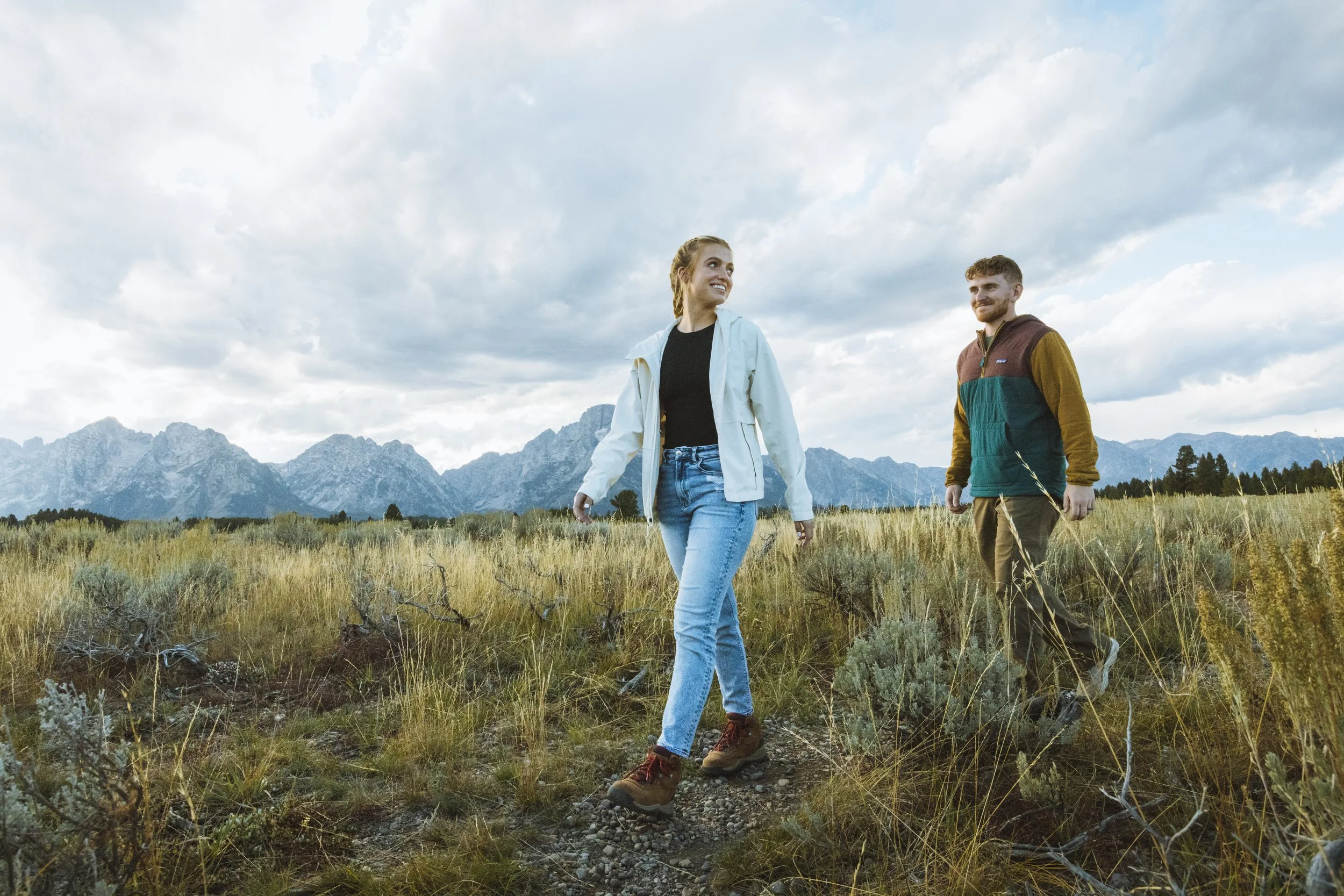 Two people hiking through a grassy field with mountain ranges in the background, under cloudy skies.