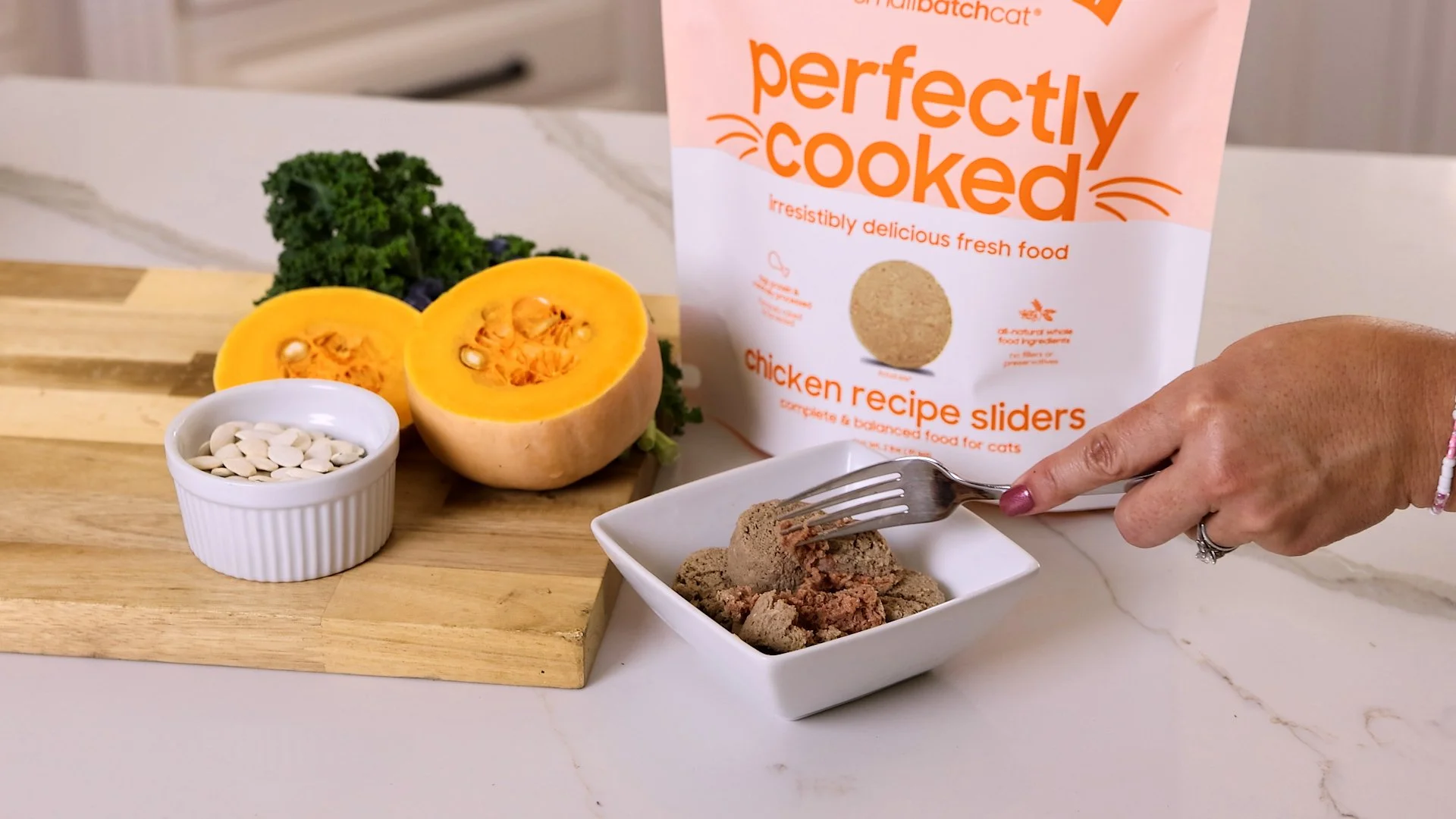 A white bowl filled with cat food chunks on a countertop, with a hand holding a fork picking up a piece. Behind, a package labeled 'perfectly cooked chicken recipe sliders' is visible. To the left, a halved butternut squash, a small white ramekin wit