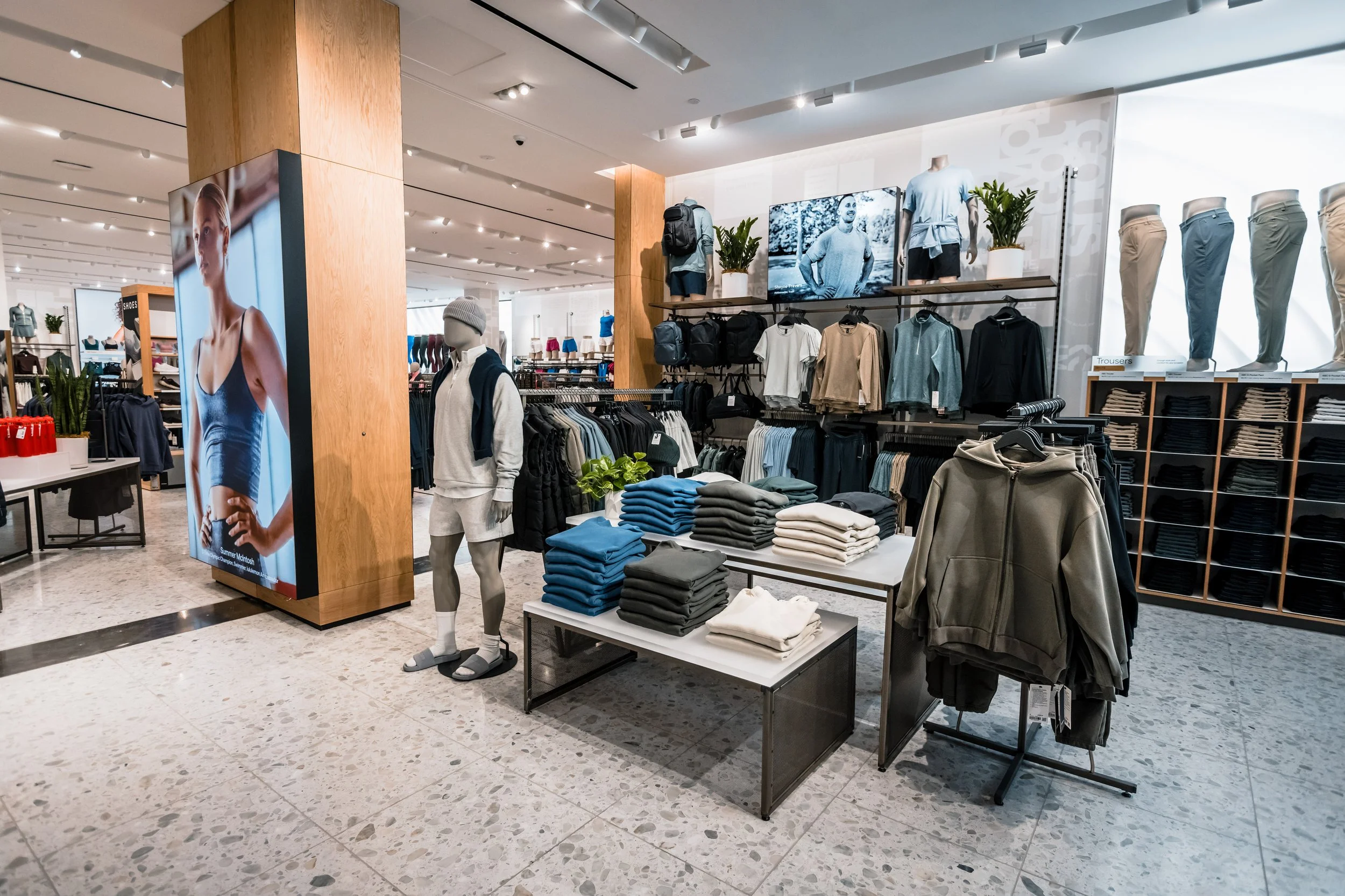 Clothing store display with folded sweatshirts, hoodies, pants, and mannequins wearing casual athletic apparel, along with digital screens showing models.